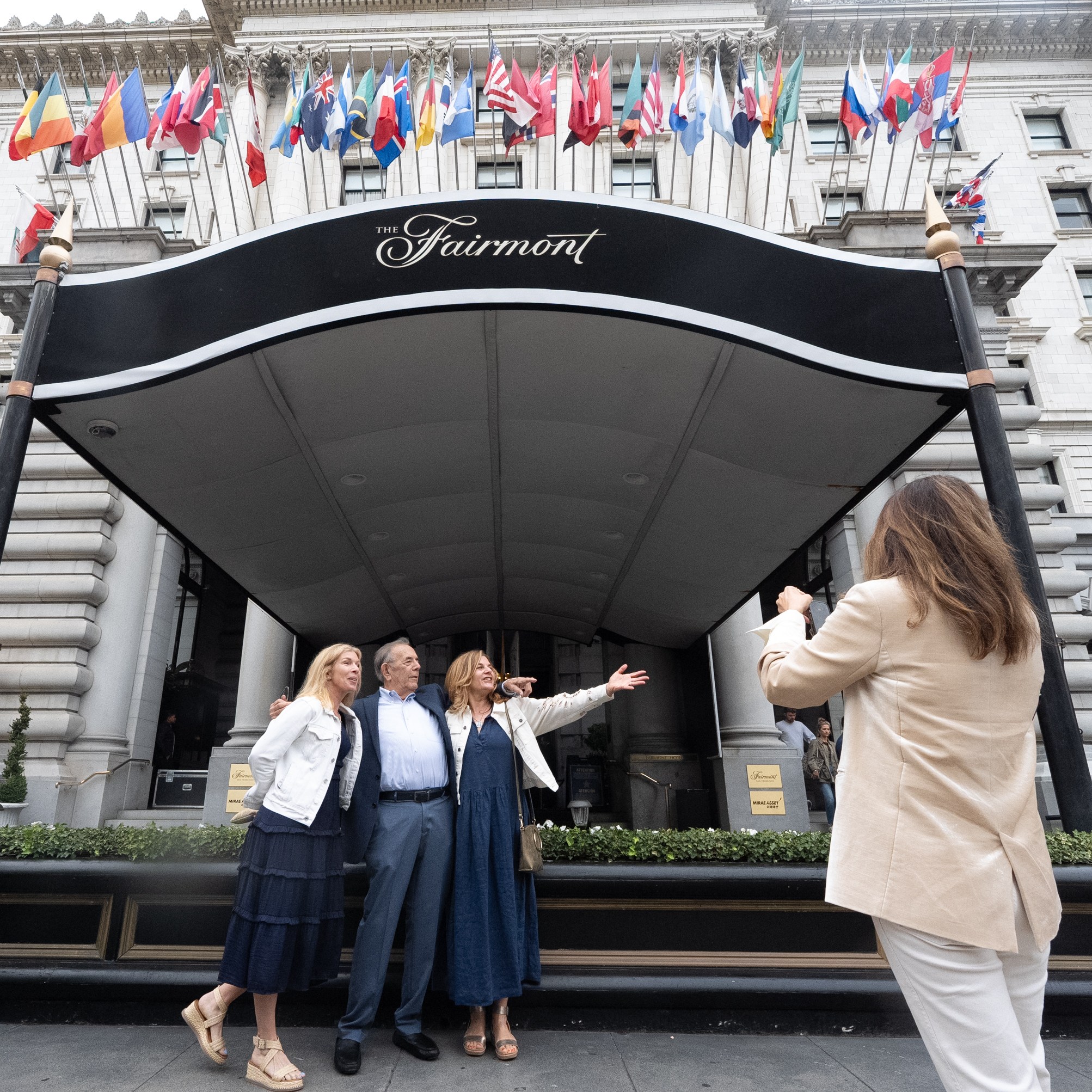 In front of a grand hotel entrance displaying international flags, three people pose for a photo while another person takes the picture, all dressed in formal-casual attire.