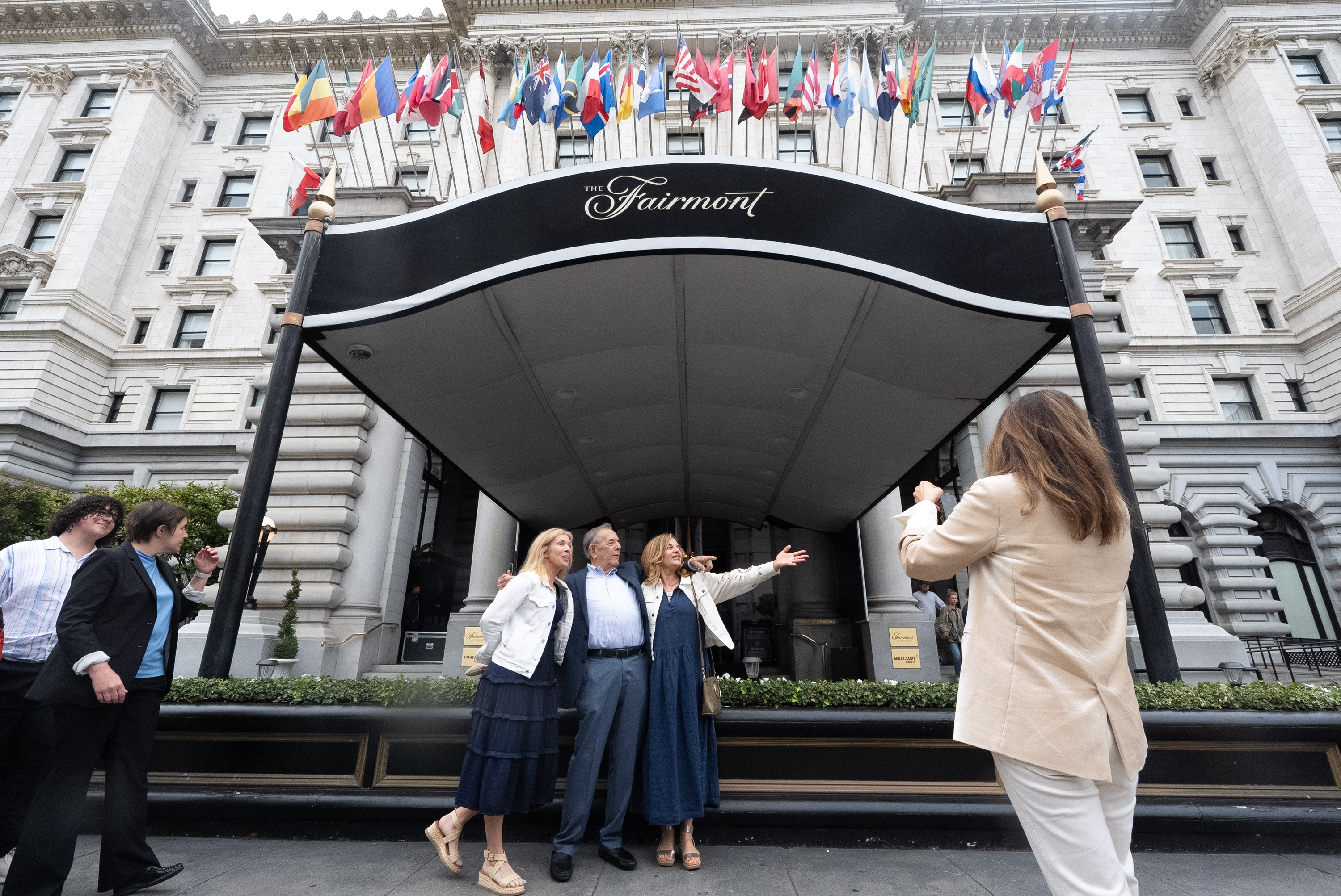 In front of a grand hotel entrance displaying international flags, three people pose for a photo while another person takes the picture, all dressed in formal-casual attire.