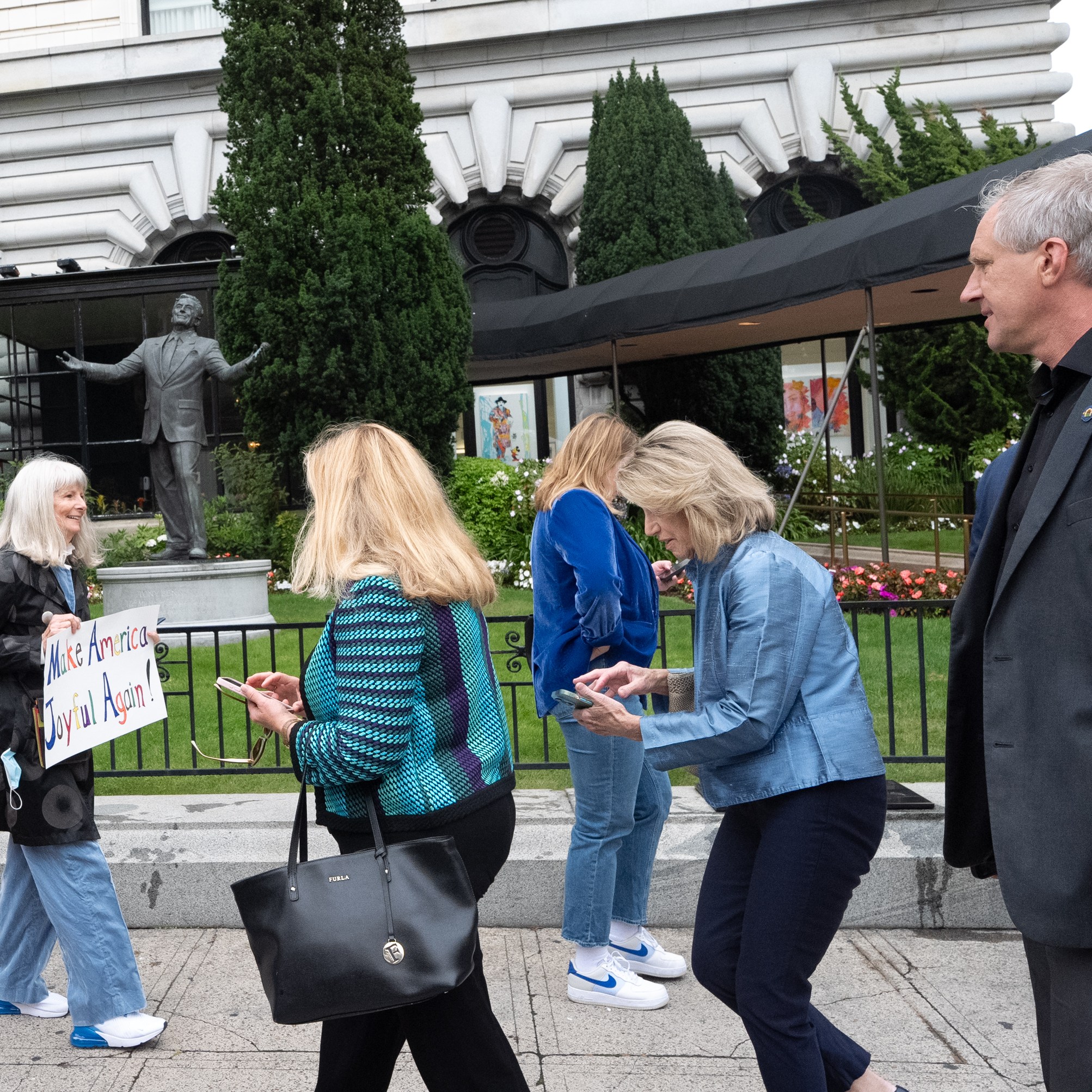 A group of people walk in front of a building, one holding a sign that reads, &quot;Make America Joyful Again!&quot; A statue with outstretched arms stands in the background.