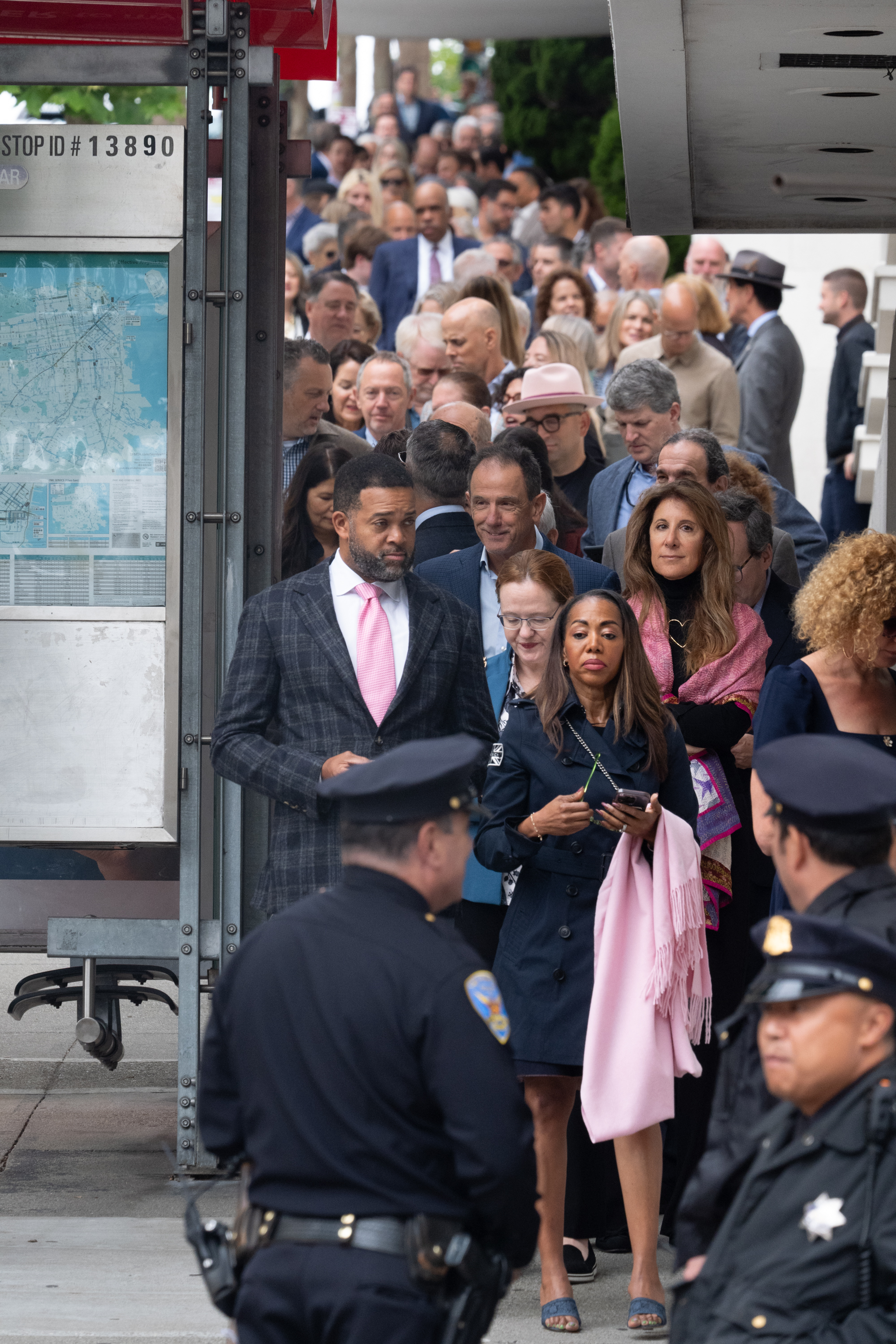 A large, diverse crowd stands in line on a sidewalk, with police officers in uniform visible in the foreground. Many people appear to be dressed formally.