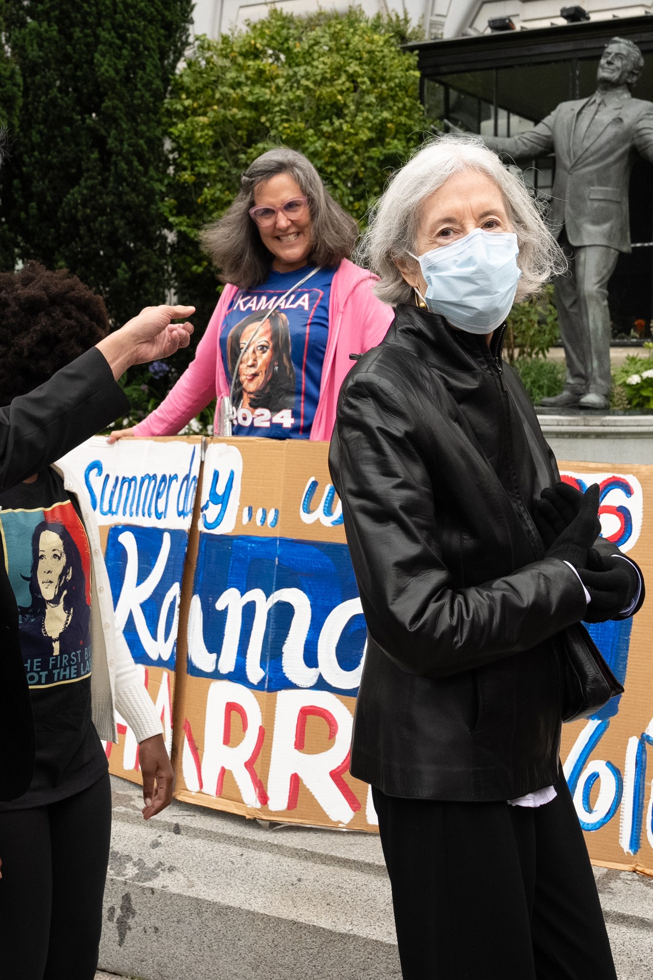 A group of people stands outside with a statue in the background, holding signs supporting Kamala Harris for 2024, with one person wearing a mask and another pointing.