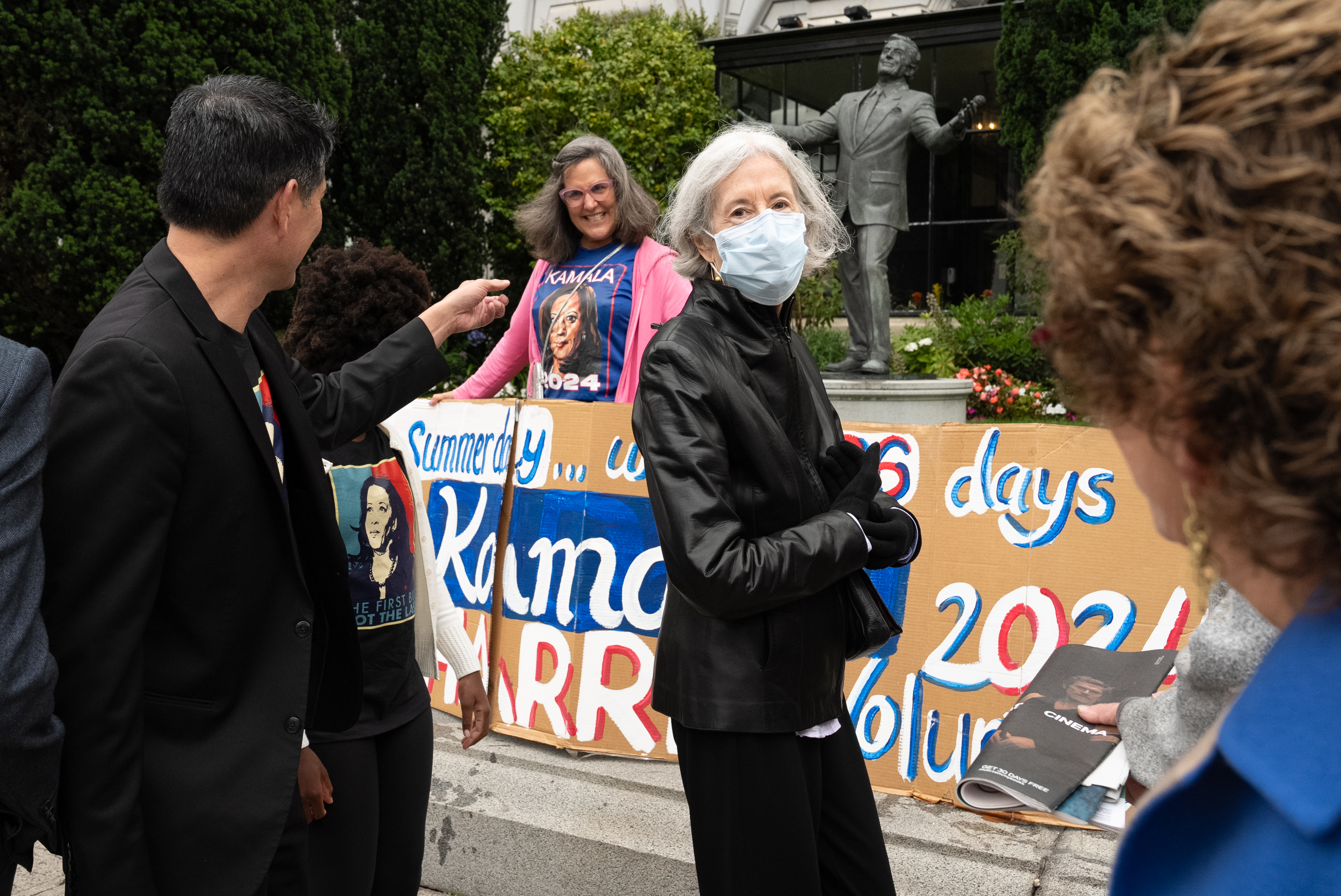 A group of people stands outside with a statue in the background, holding signs supporting Kamala Harris for 2024, with one person wearing a mask and another pointing.