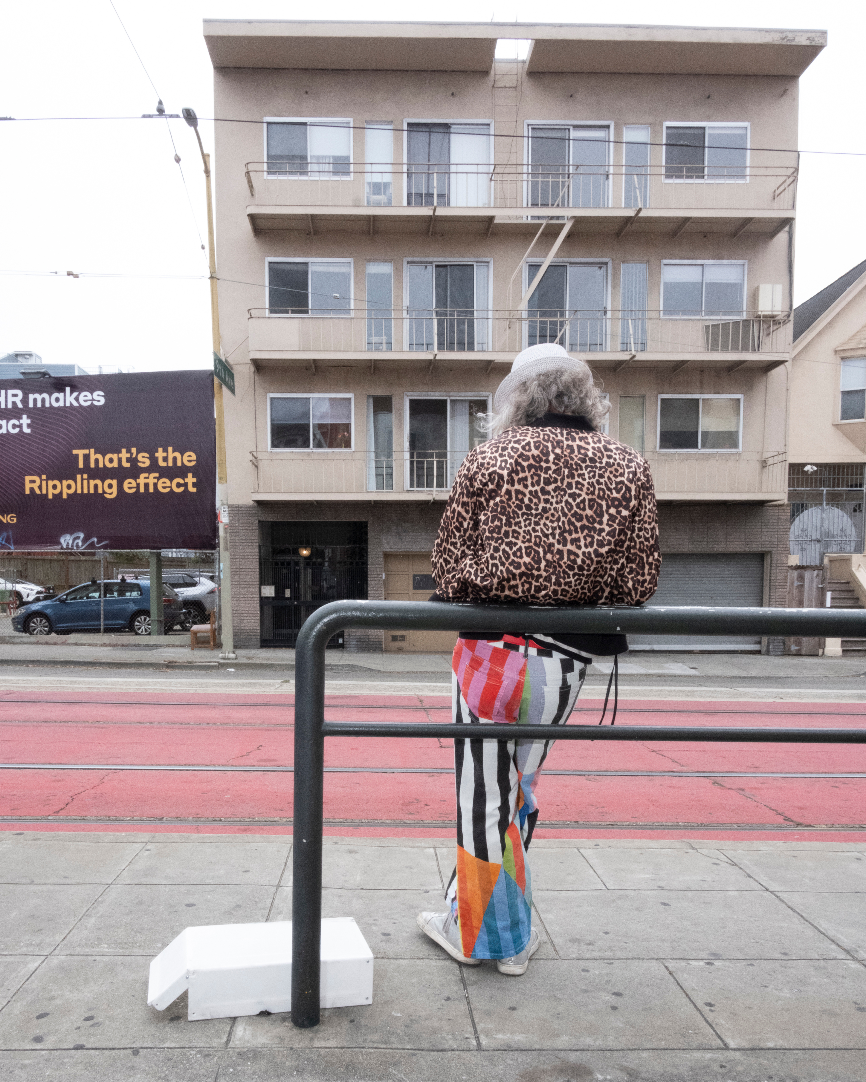 A person wearing a leopard print jacket, colorful striped pants, and a white hat stands facing an apartment building, with a street and billboard to the left.