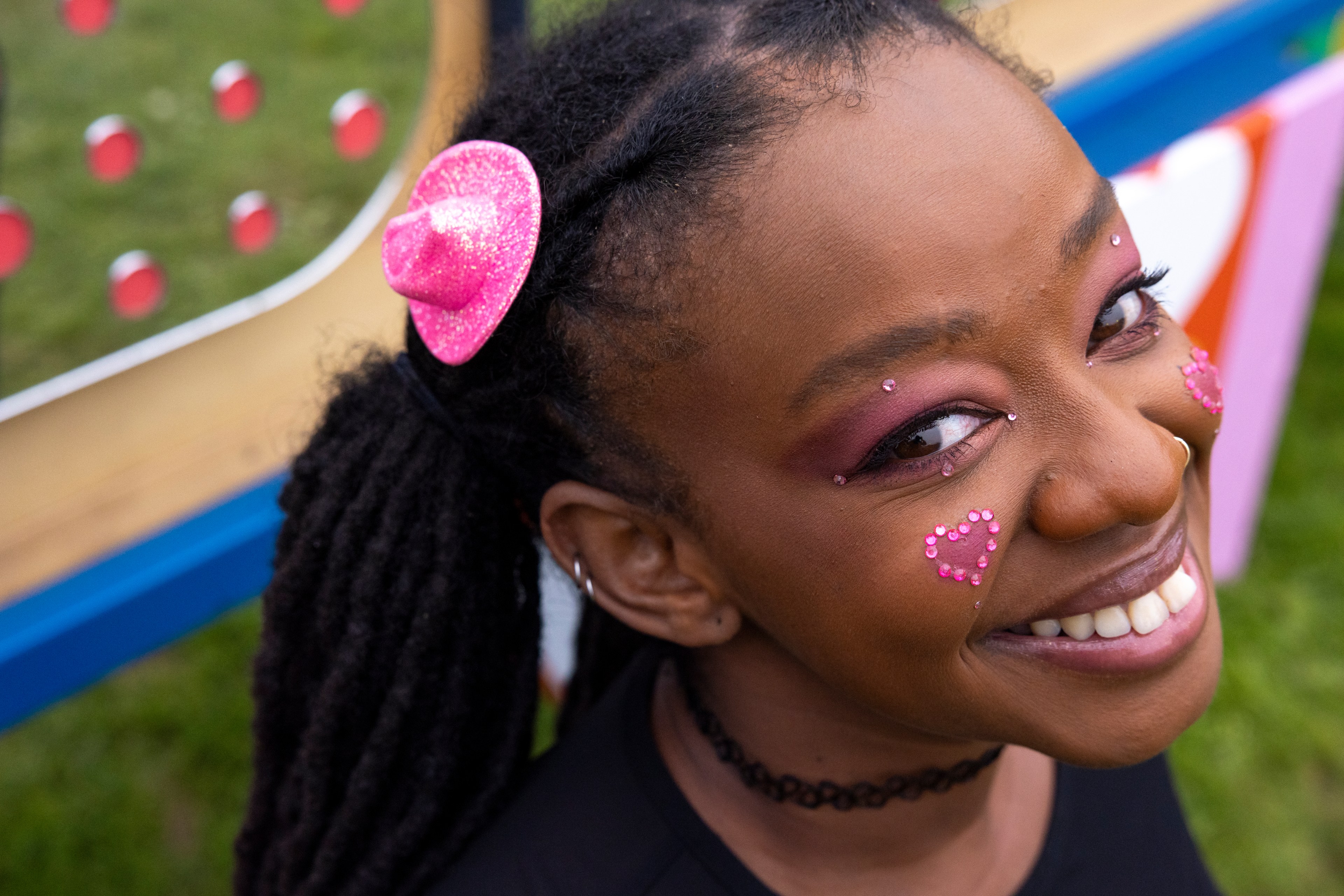 A smiling person with a pink glittery hat accessory, pink eye makeup, and heart-shaped face gems. The background shows an outdoor setting with colorful, blurred objects.