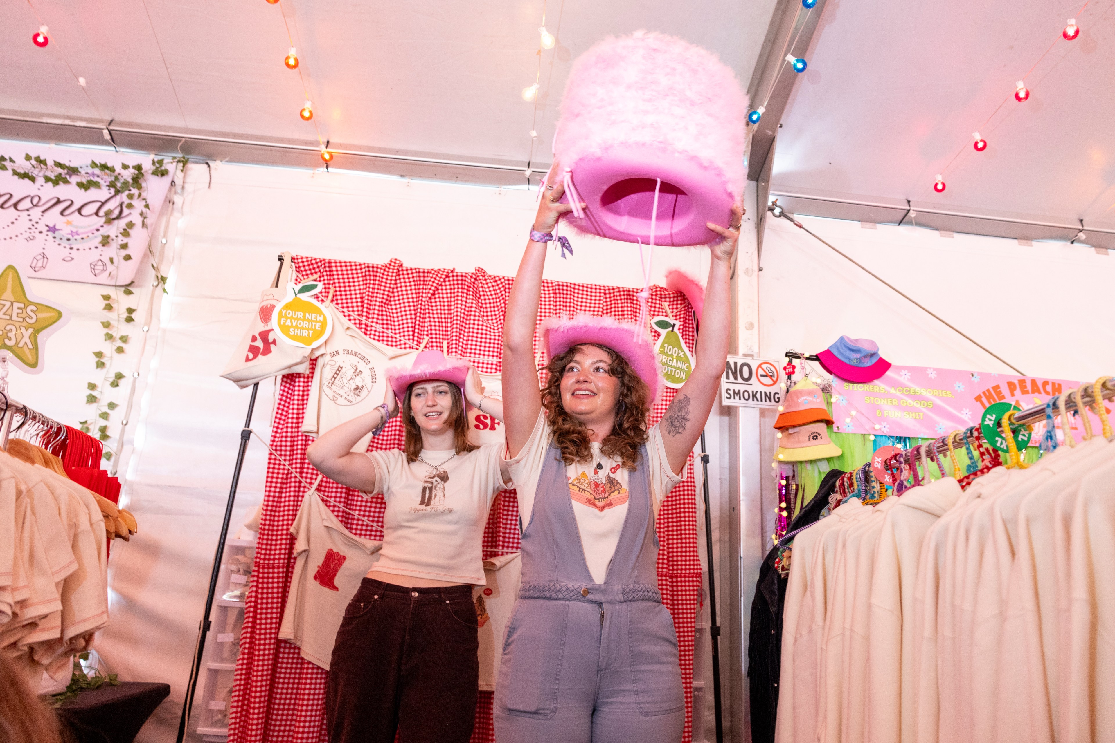 Two people are inside a vibrant, decorated booth. They are smiling and wearing pink cowboy hats, with one person lifting a large pink fluffy hat above their head.