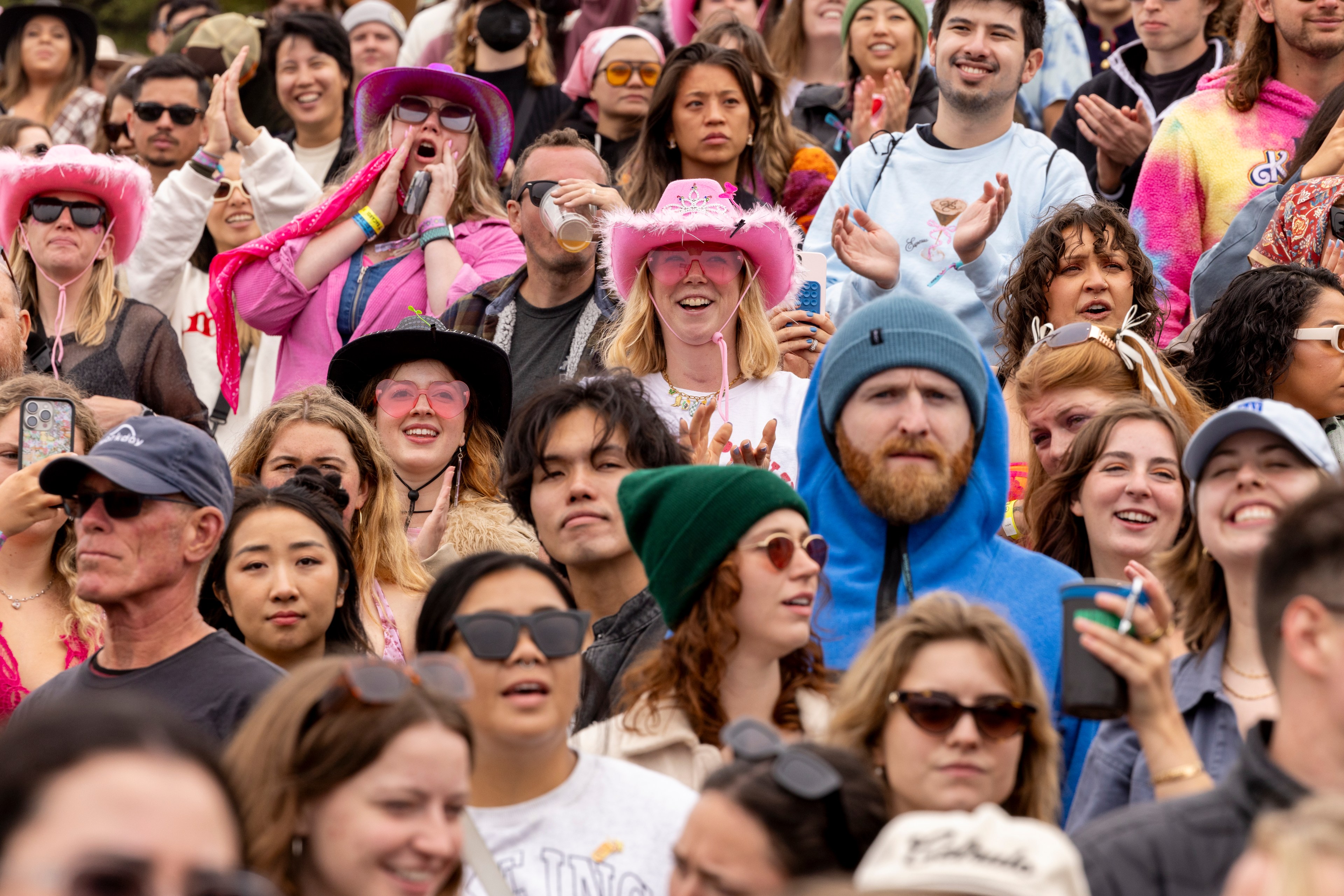 A lively crowd of people, many wearing colorful hats and sunglasses, are gathered outdoors, smiling and clapping, enjoying a festive event.