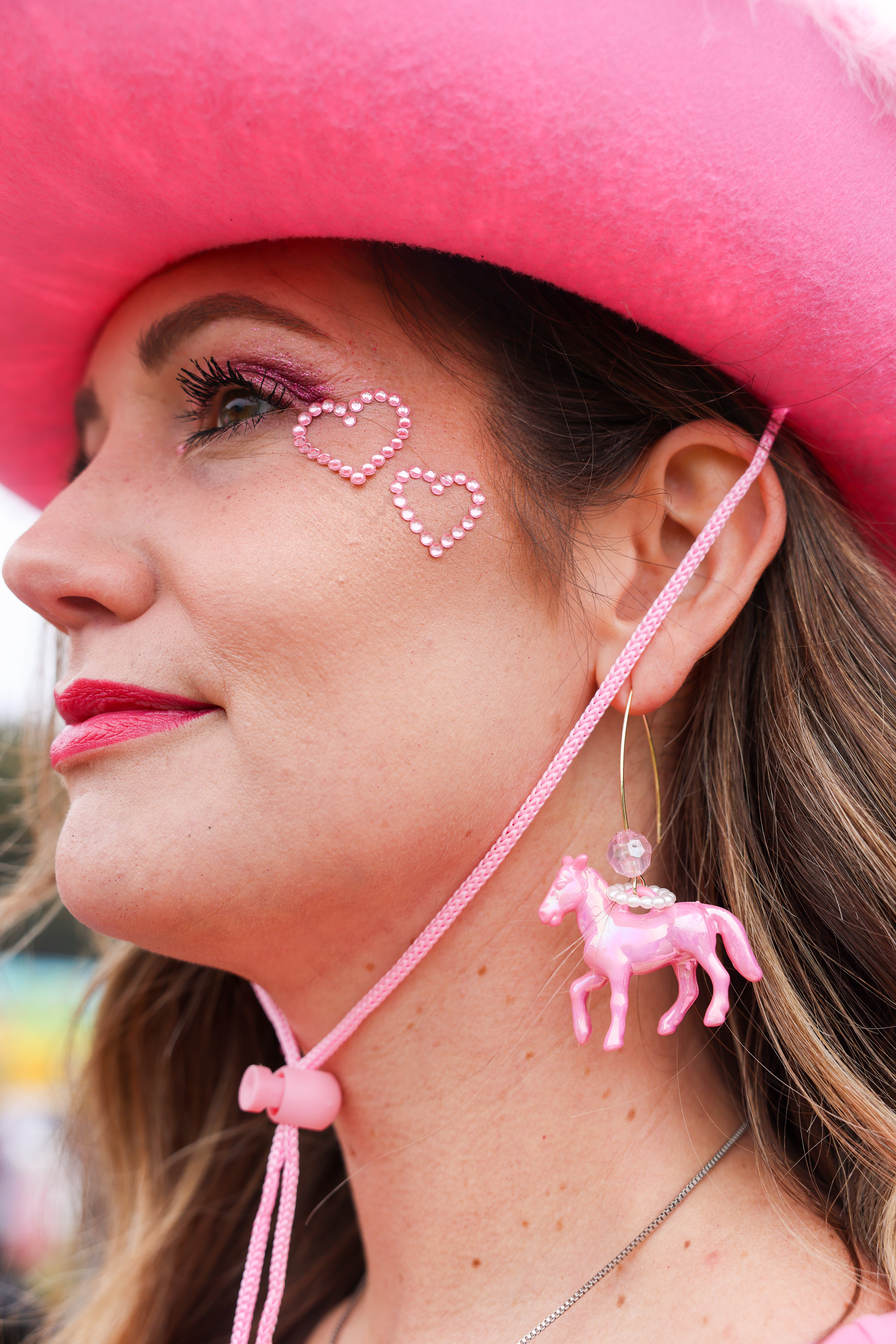 A woman is seen wearing a bright pink cowboy hat, with pink heart rhinestones by her eyes, and a pink horse-shaped earring. She has pink lipstick and detailed eye makeup.