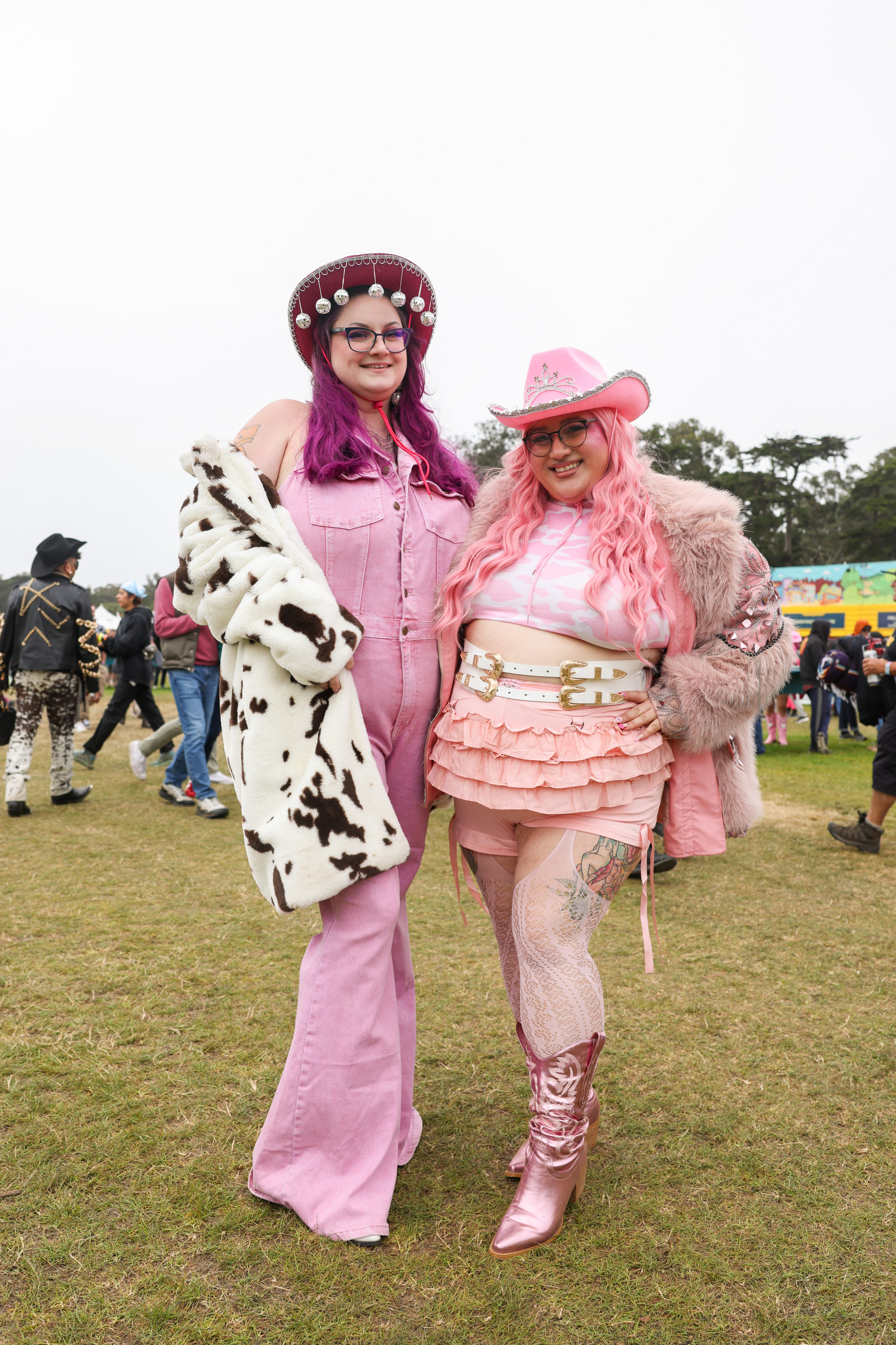 Two people dressed in vibrant pink outfits stand outdoors on a grassy area. One wears a pink jumpsuit and cow-print coat, the other a pink ruffled outfit with cowboy boots.