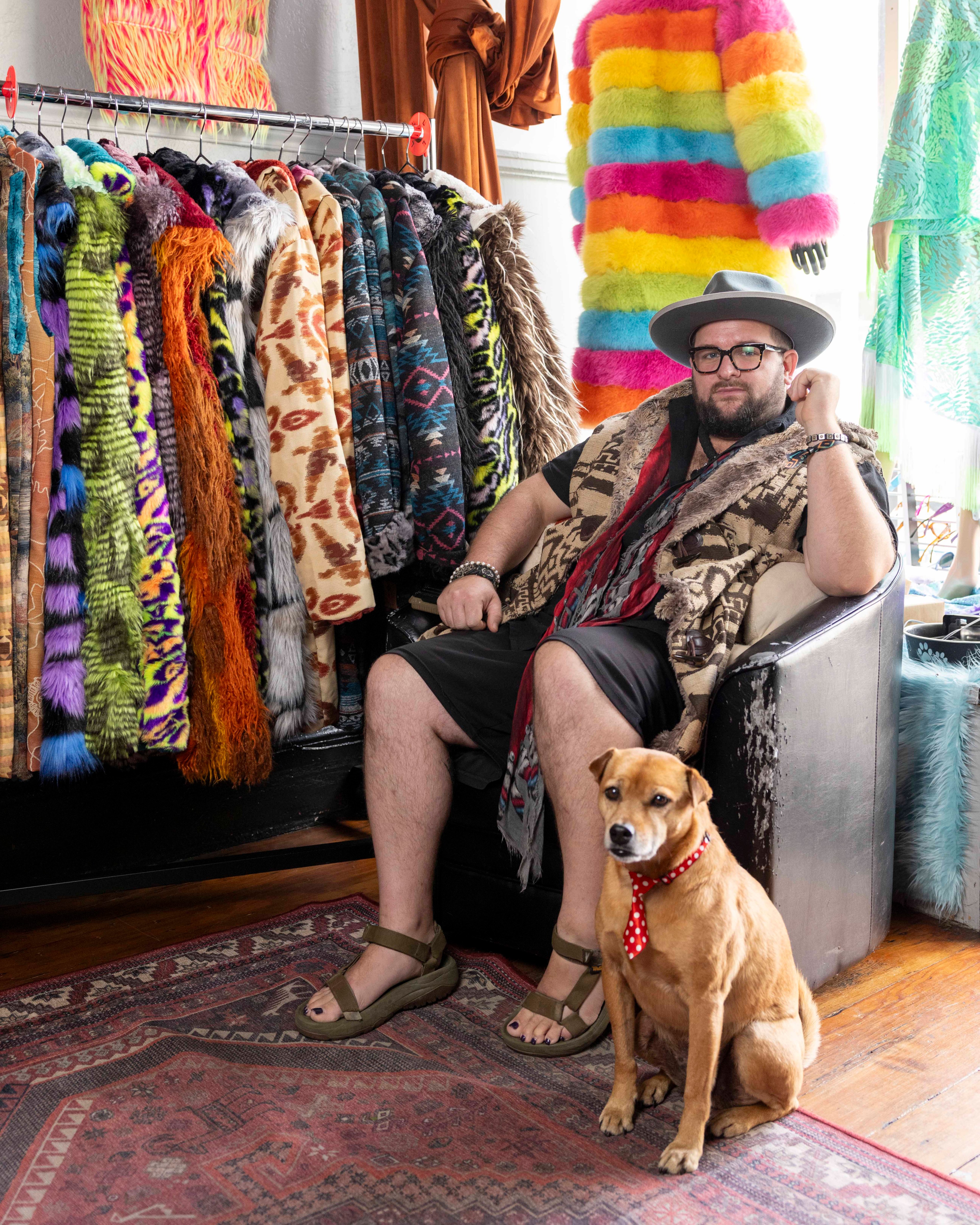 A bearded man in a hat and sandals sits in a chair next to a dog with a red polka dot bandana, surrounded by colorful fur coats and outfits on racks and mannequins.