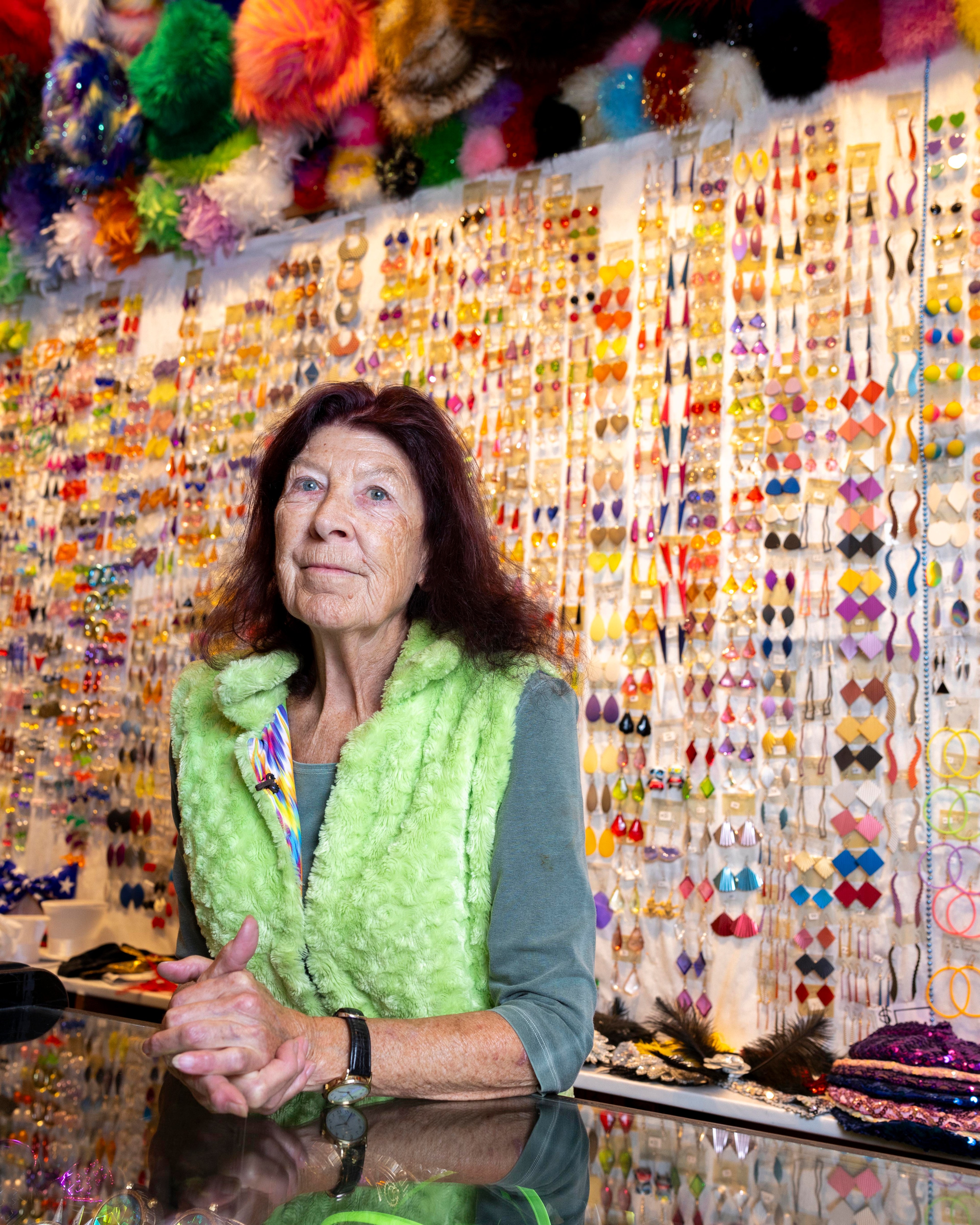 An elderly woman is standing behind a counter in front of a wall filled with colorful hanging earrings. She wears a lime green fuzzy vest and has variegated items above and around her.