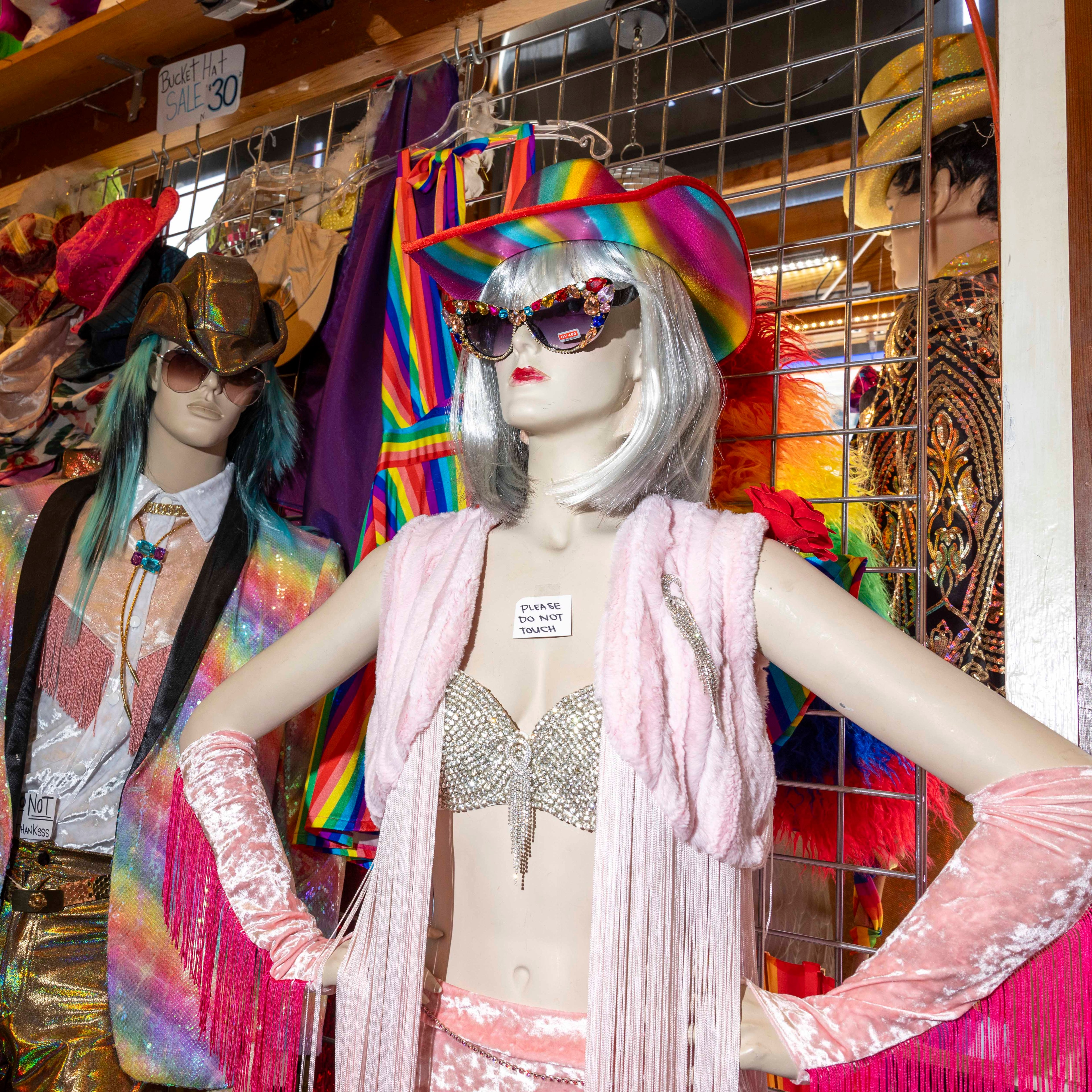 Two mannequins in colorful, eccentric outfits stand in a vibrant shop. One wears a rainbow hat, sparkly bra, and sunglasses; the other has metallic clothes and green hair.