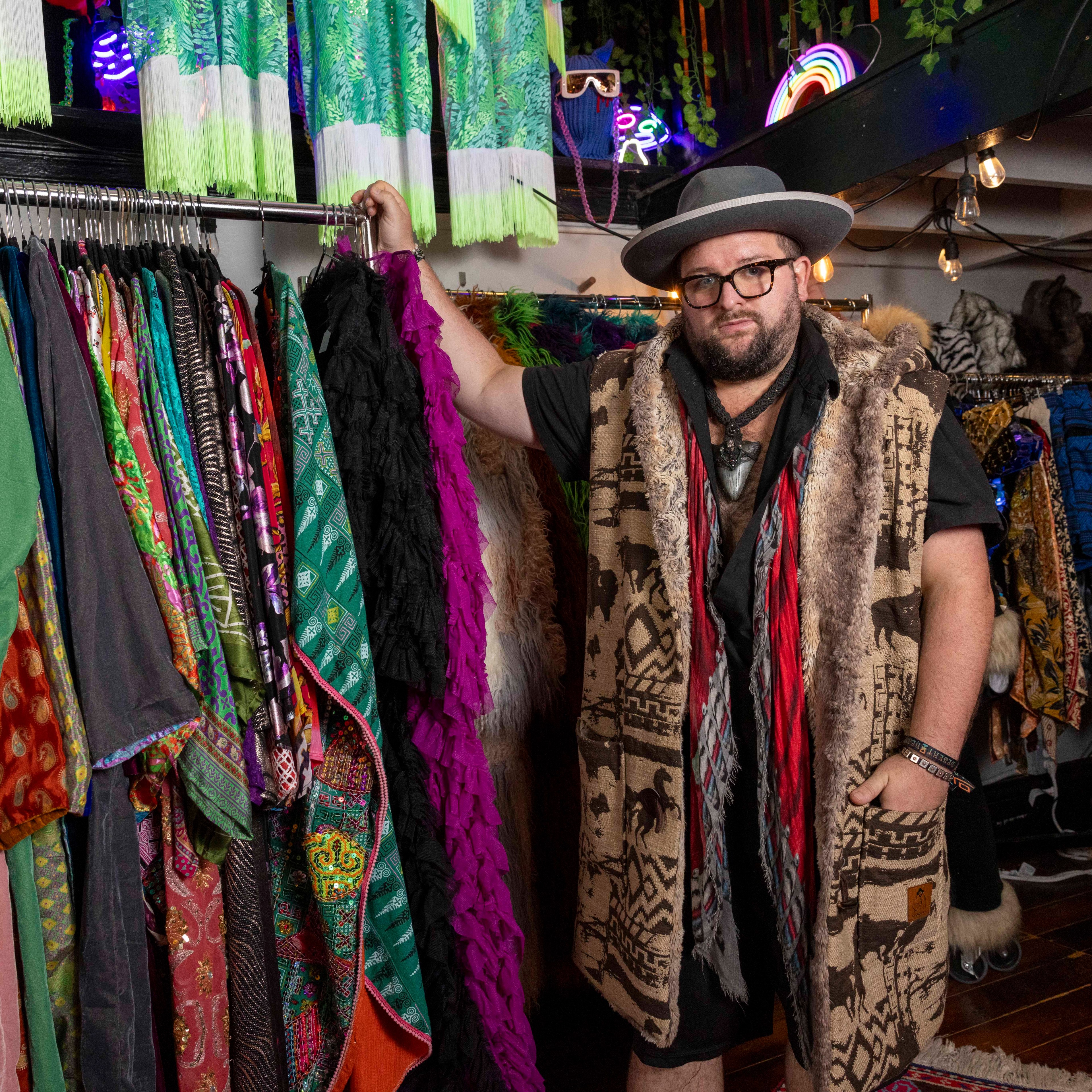 A man wearing a wide-brimmed hat and a patterned fur-trimmed vest stands in a colorful clothing store, holding a black ruffled garment and surrounded by vibrant fabrics.