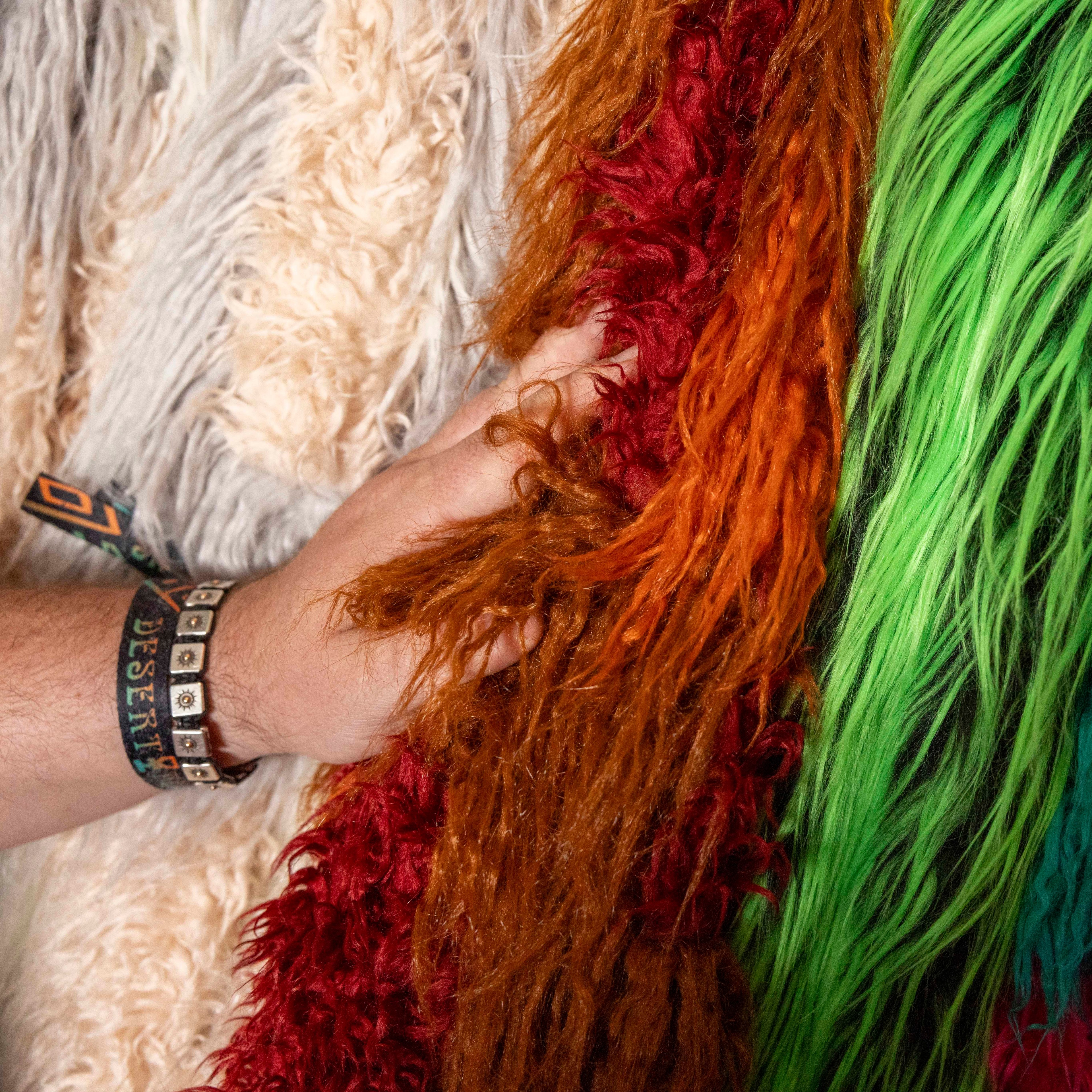 A hand is grasping fuzzy, textured fabrics in various vibrant colors, including green, red, orange, and cream, with the person wearing bracelets on their wrist.