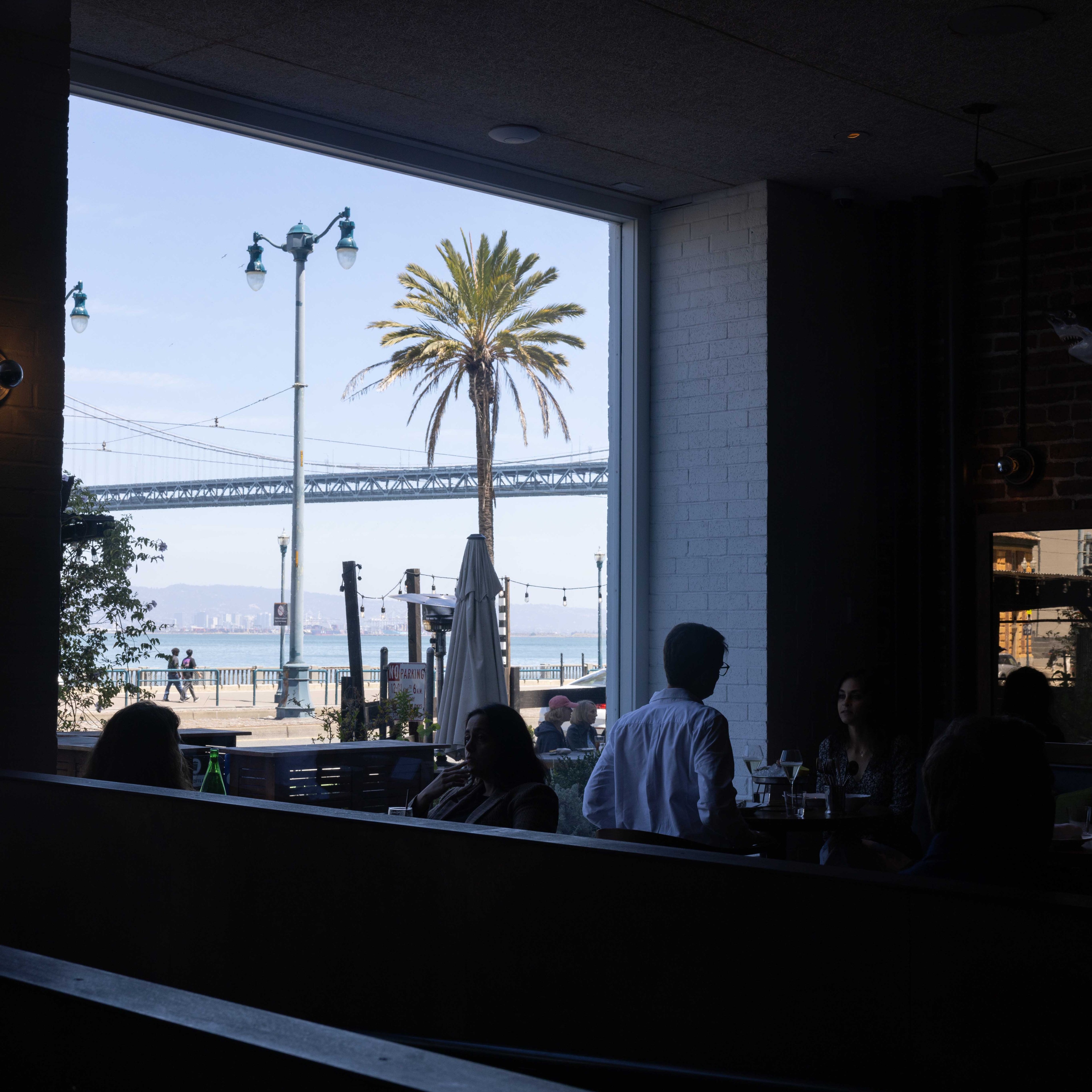 The image shows people dining inside a dimly-lit restaurant with a large window framing an outdoor view of a street, a palm tree, and a bridge over water.