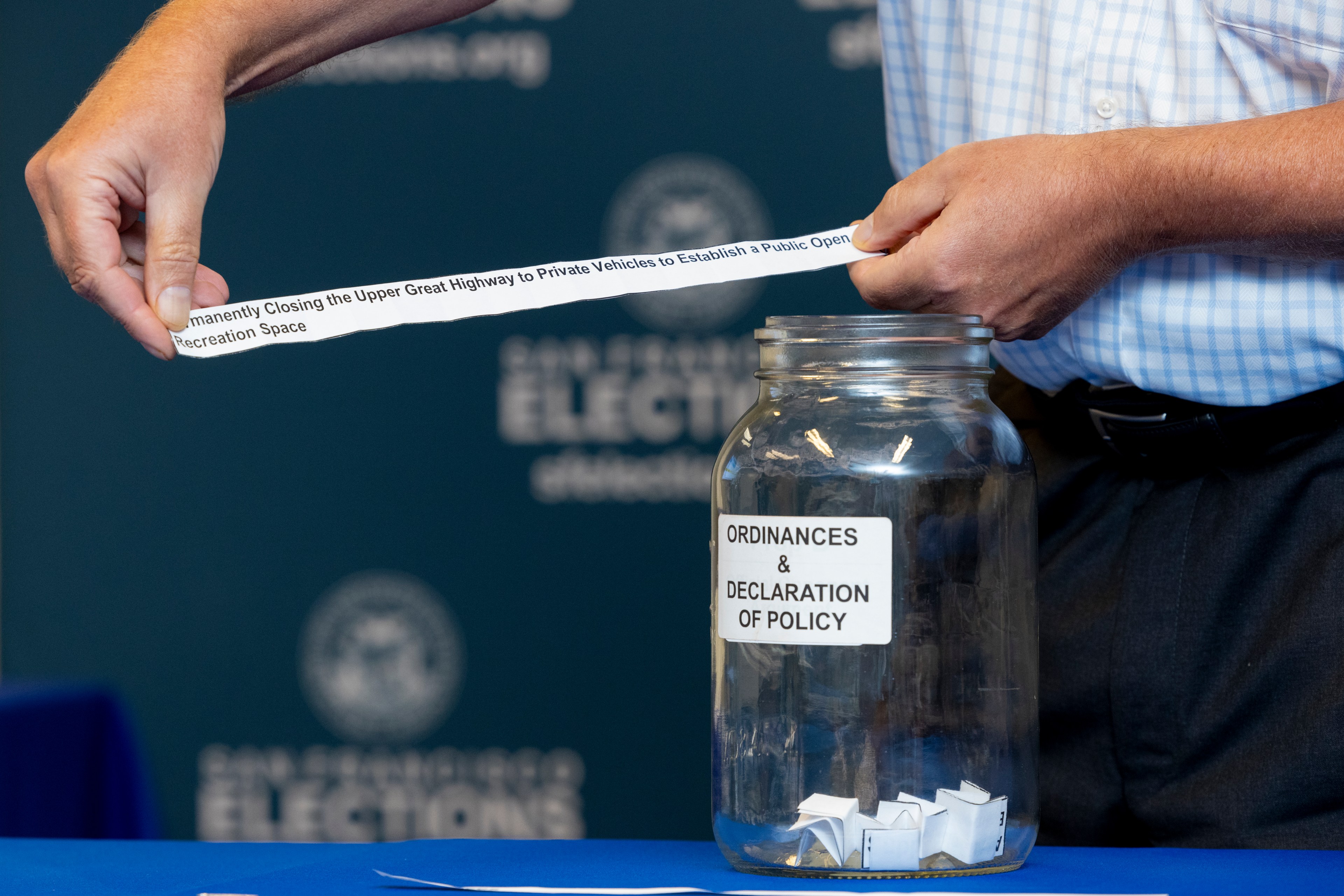 Two hands pull a strip of paper reading &quot;Permanently Closing the Upper Great Highway to Private Vehicles...&quot; from a glass jar labeled &quot;Ordinances &amp; Declaration of Policy.&quot;