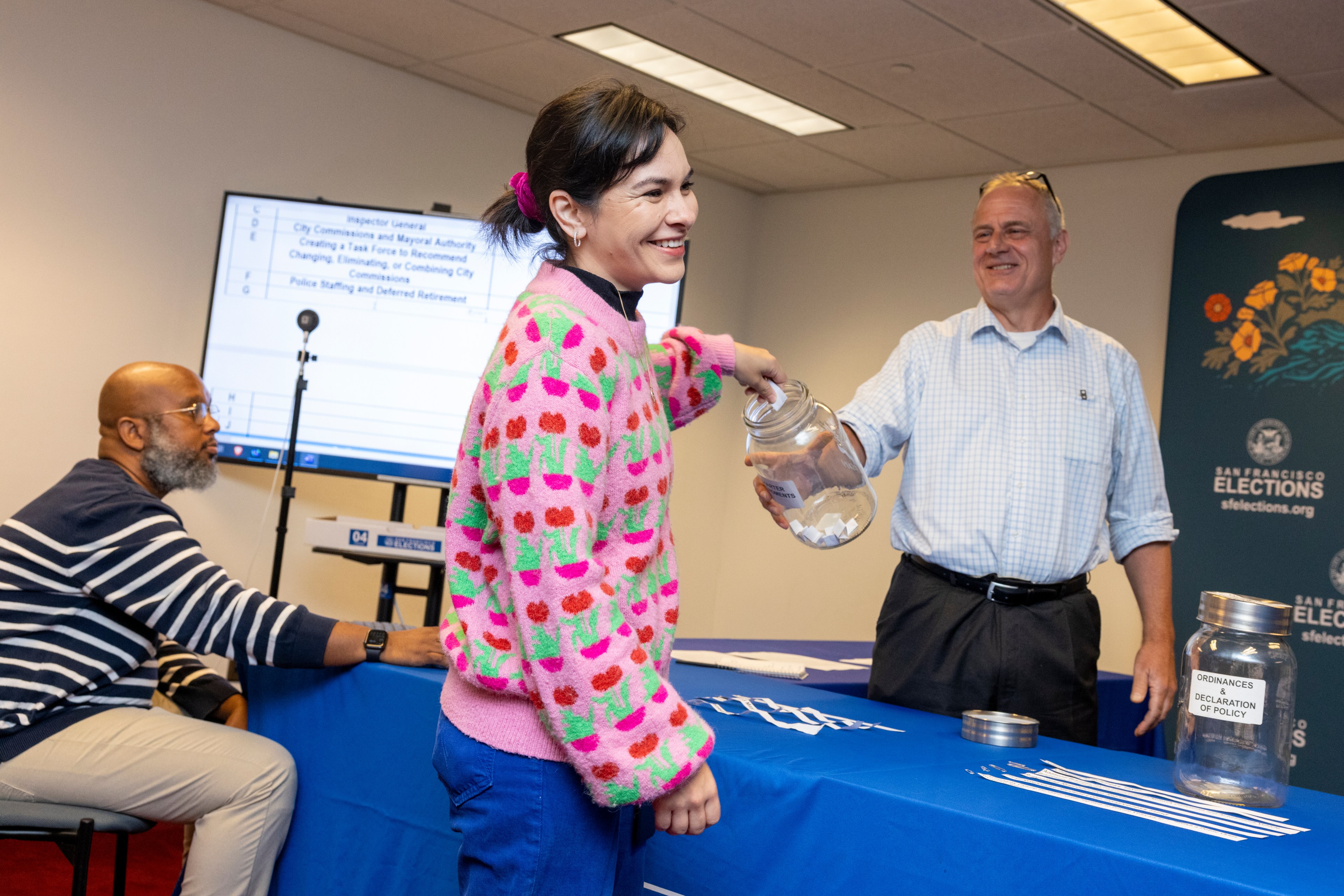 A smiling woman in a pink sweater draws items from a jar at a table, while two men nearby look on, one seated and one standing, in an election event setting.