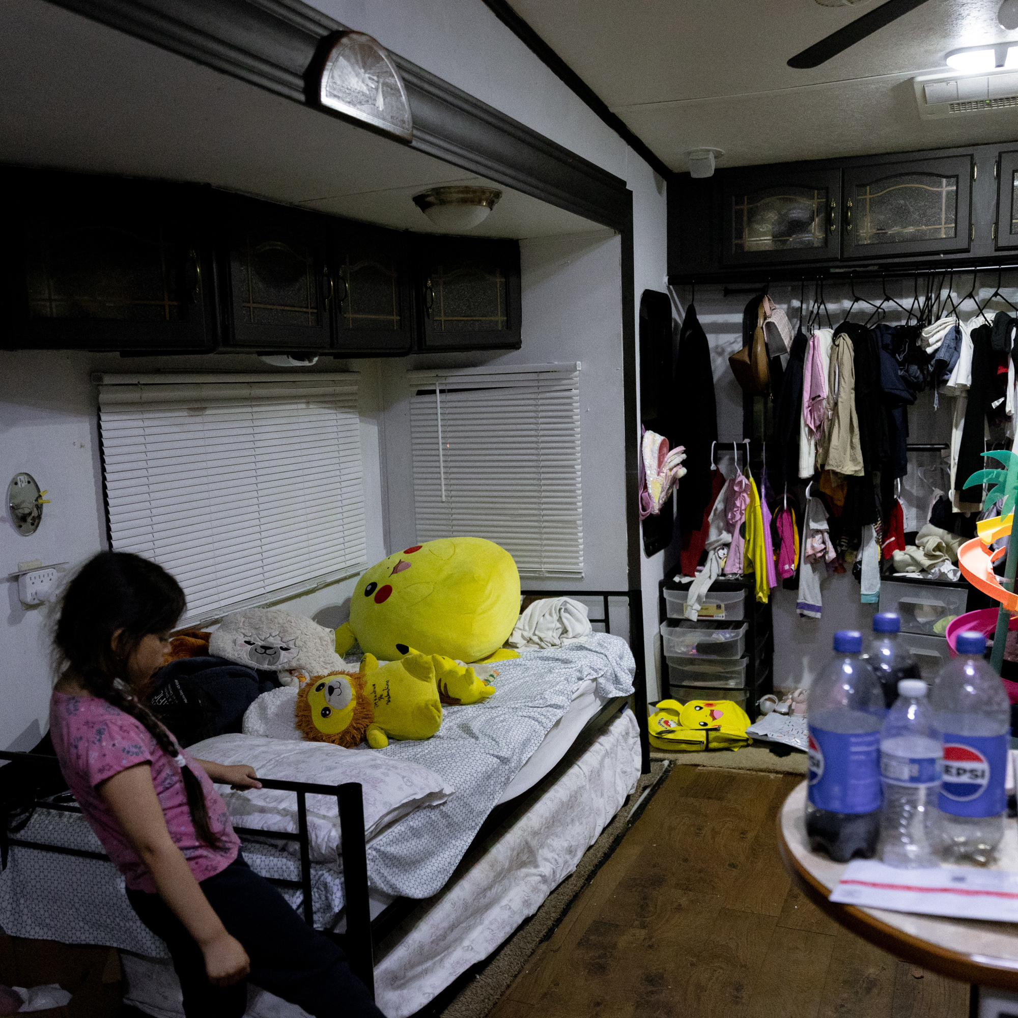 A cluttered room with a girl standing by a bed that has large plush toys. On the right, clothes hang while a table holds water and soda bottles. The setting appears lived-in.