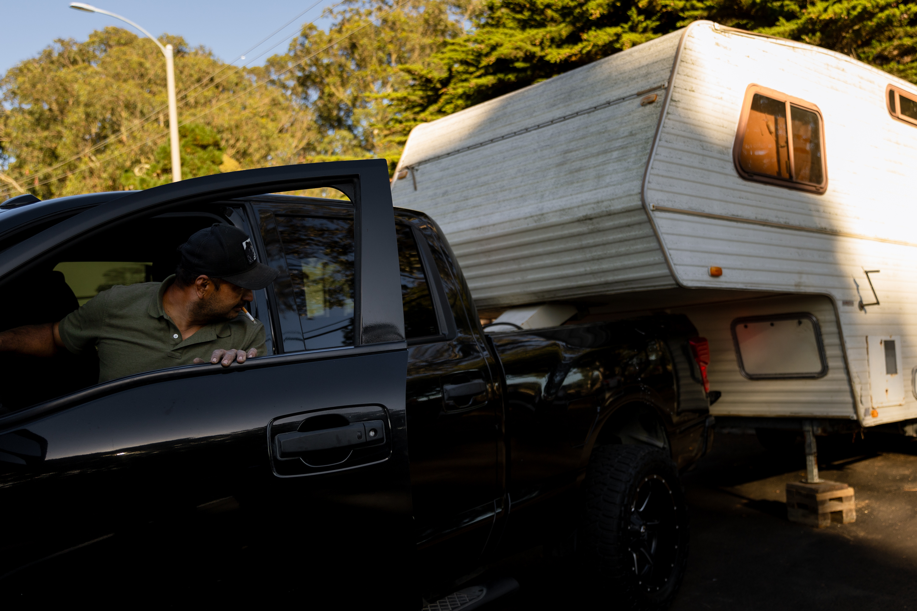 A man in a black cap and green shirt is stepping out of a black pickup truck, which is parked next to a white camper trailer. Trees are visible in the background.