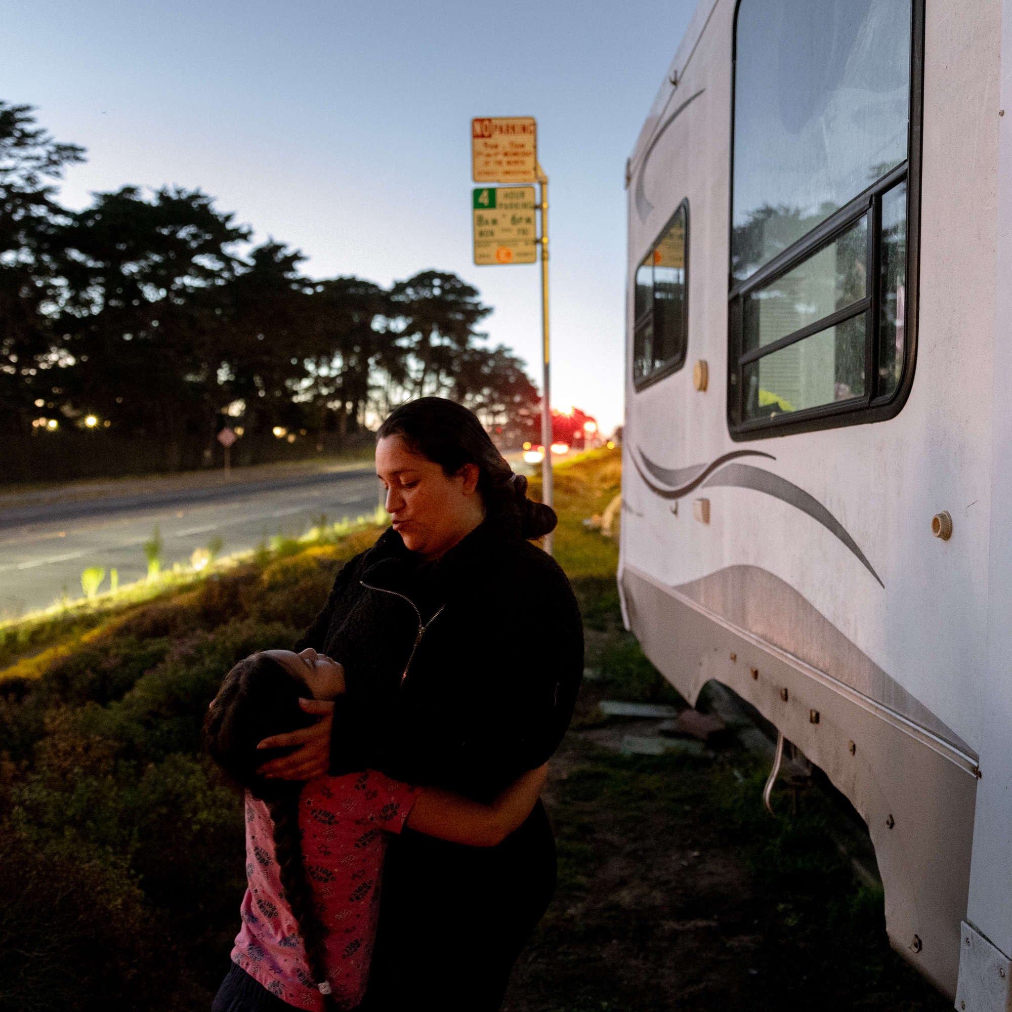 A woman and a child share an embrace near a parked RV along a road at dusk, with trees and a &quot;No Parking&quot; sign in the background.