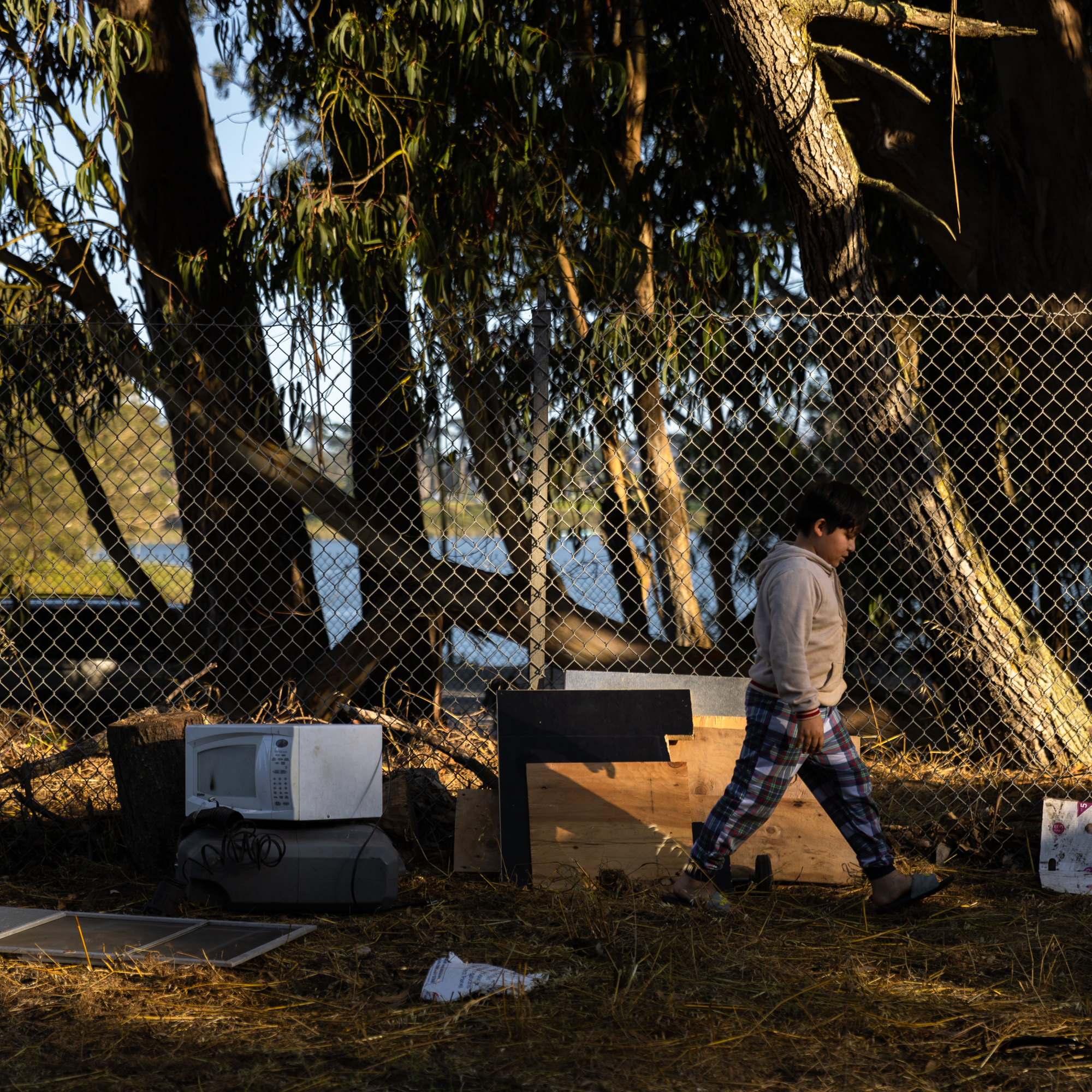 A child in a gray hoodie and checkered pants walks by a chain-link fence in a wooded area with scattered old appliances and wooden planks on the ground.