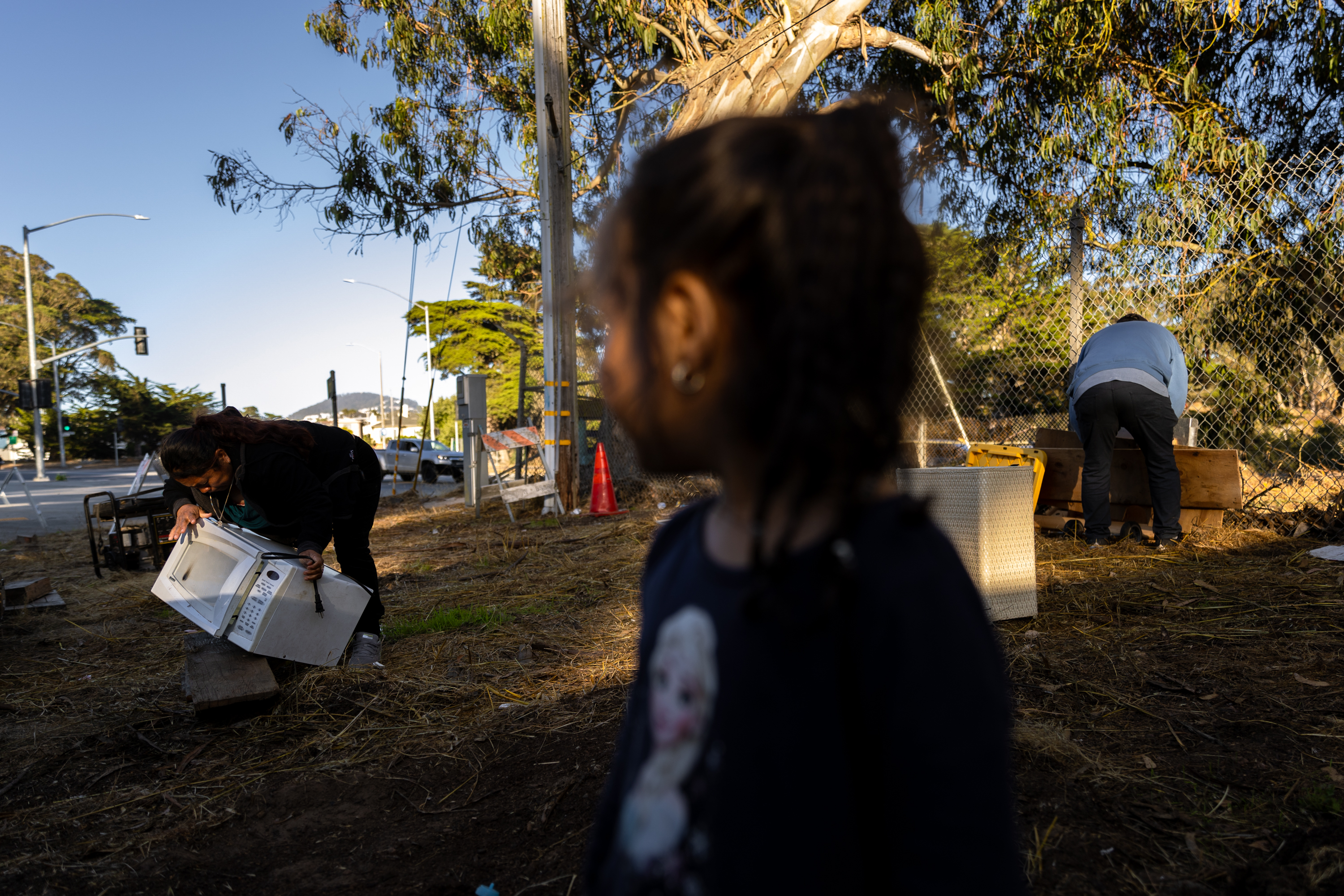 A child with braids faces away; two adults work nearby, one lifting a microwave beside a road and fence, with trees and road signs visible in the background.