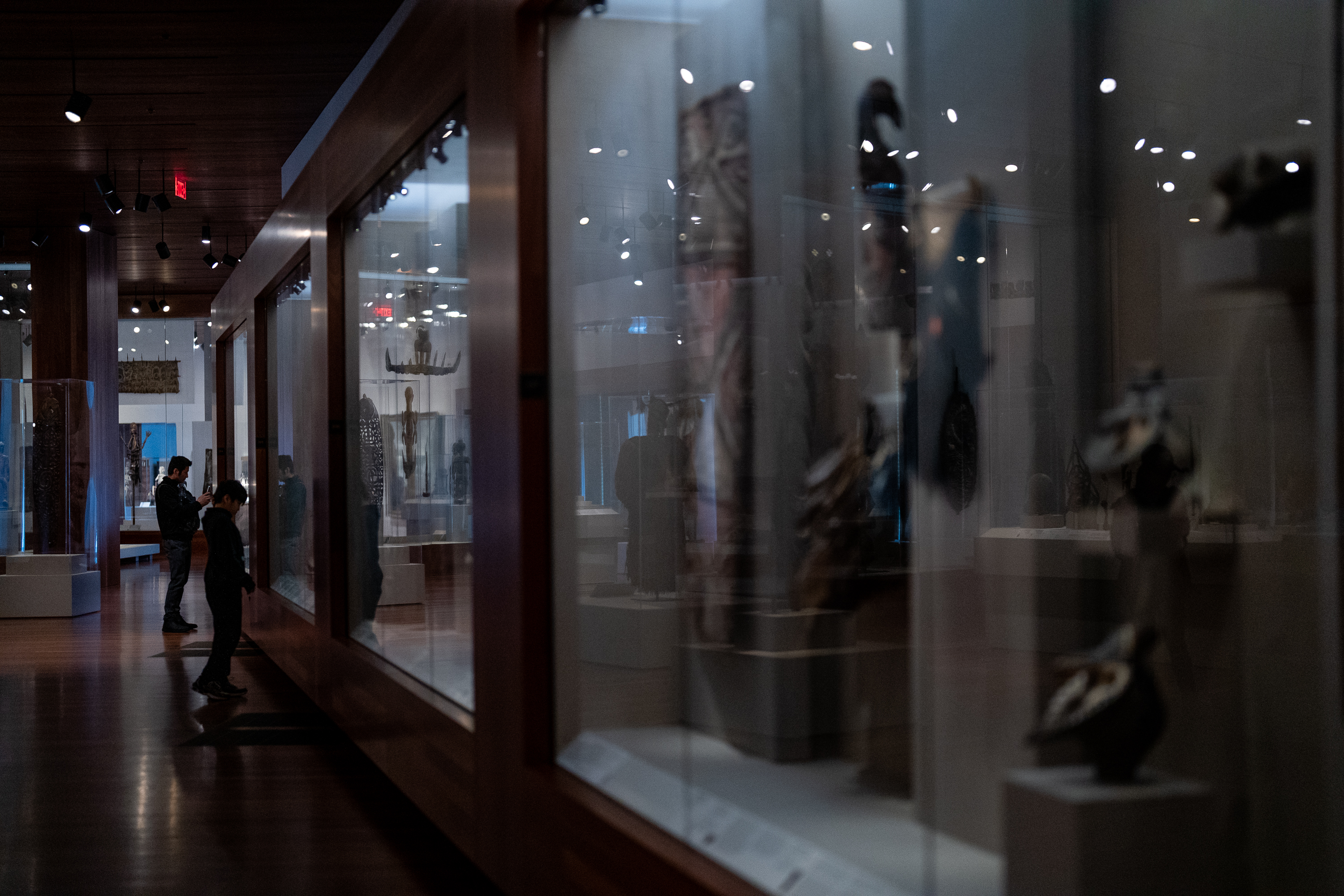 The image shows a dimly lit museum gallery with glass display cases containing artifacts. Two people are viewing the displays, one taking a photo, the other examining items closely.