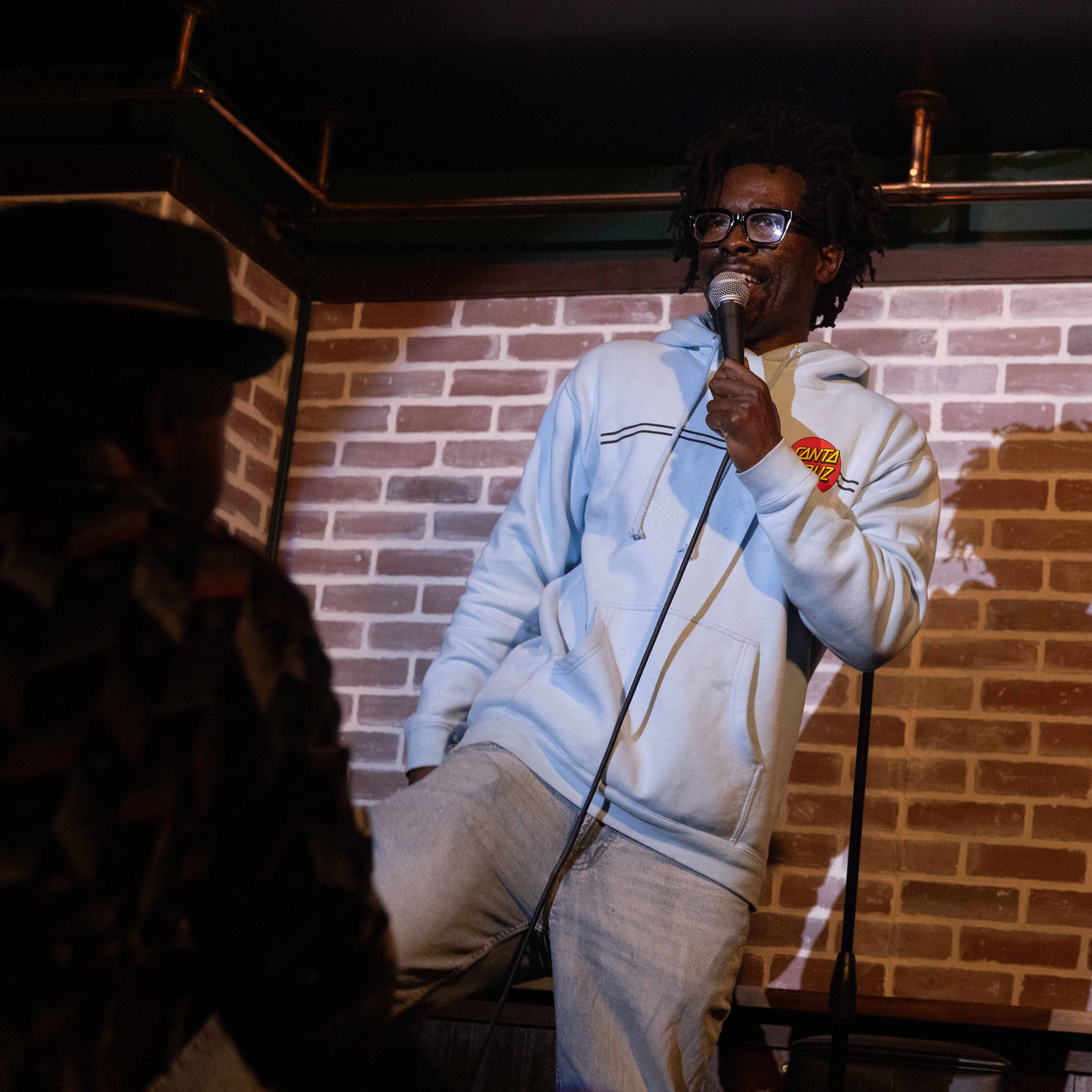 A man in a light blue hoodie and glasses is holding a microphone and performing in front of a brick wall. He stands casually, engaging with the audience.