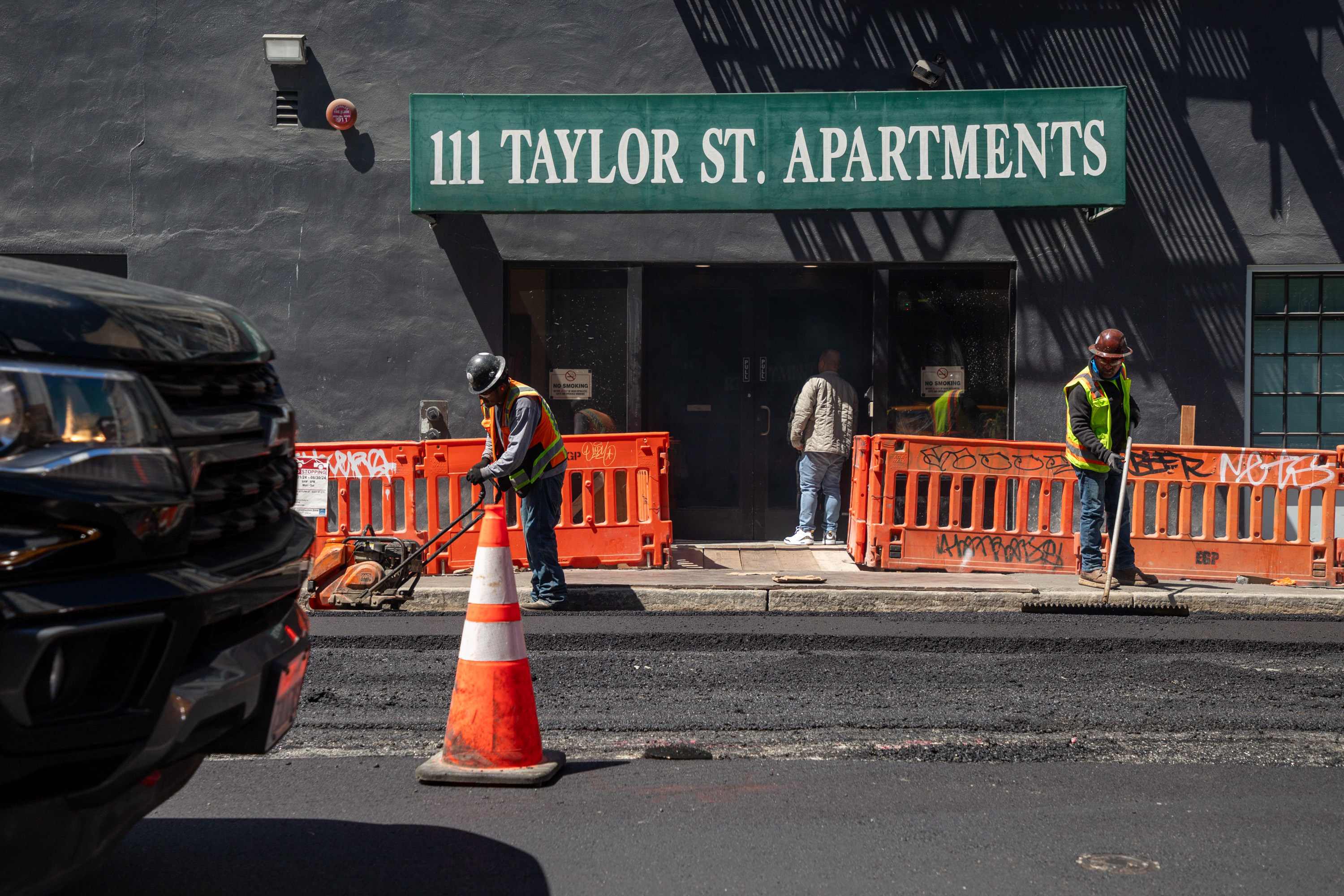 Two construction workers in safety vests and helmets work on road repairs outside &quot;111 Taylor St. Apartments,&quot; with orange barriers and a traffic cone nearby.