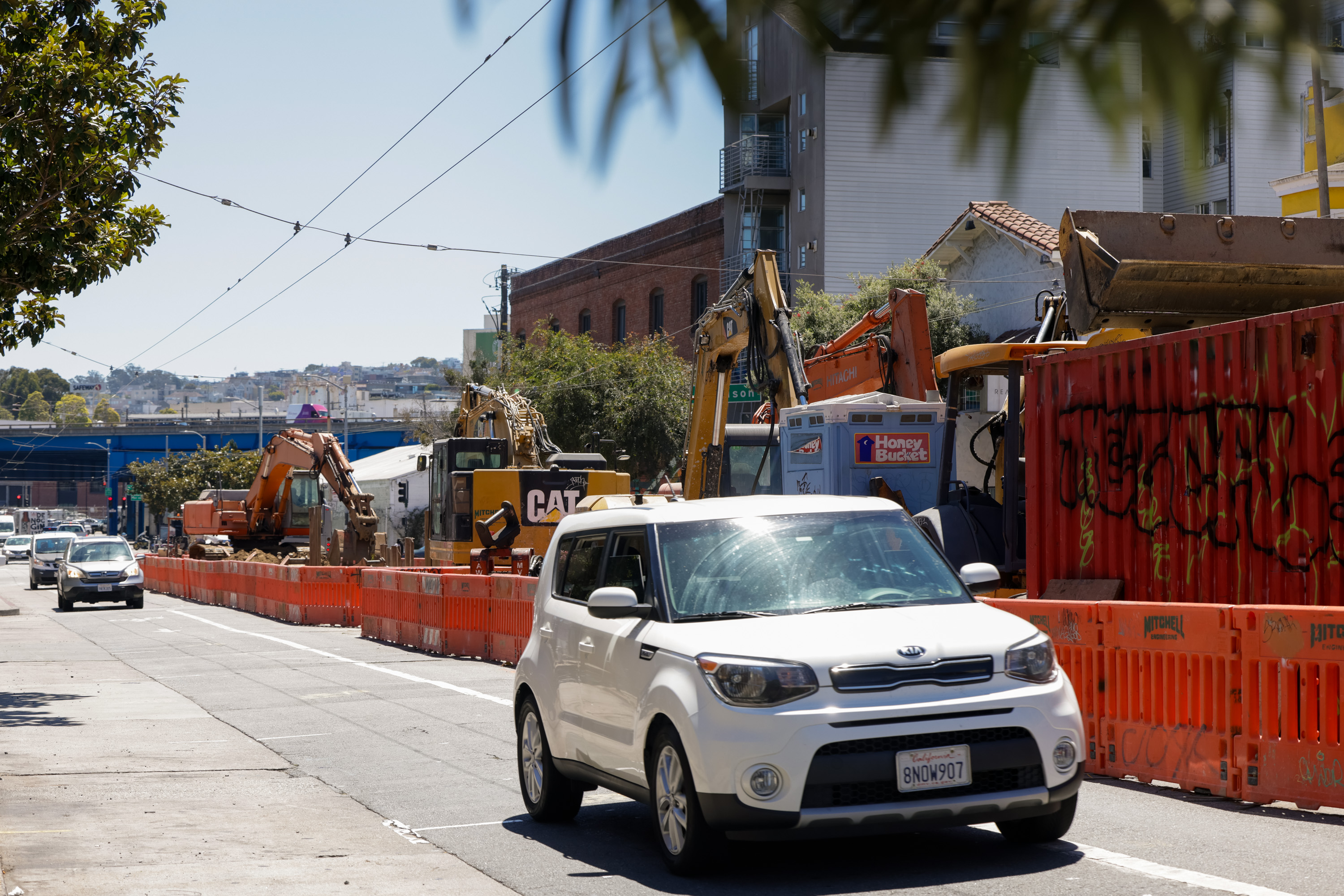 A white car drives past a construction site with orange barriers, excavators, and a &quot;Honey Bucket&quot; portable toilet, on a sunny day with a clear sky.