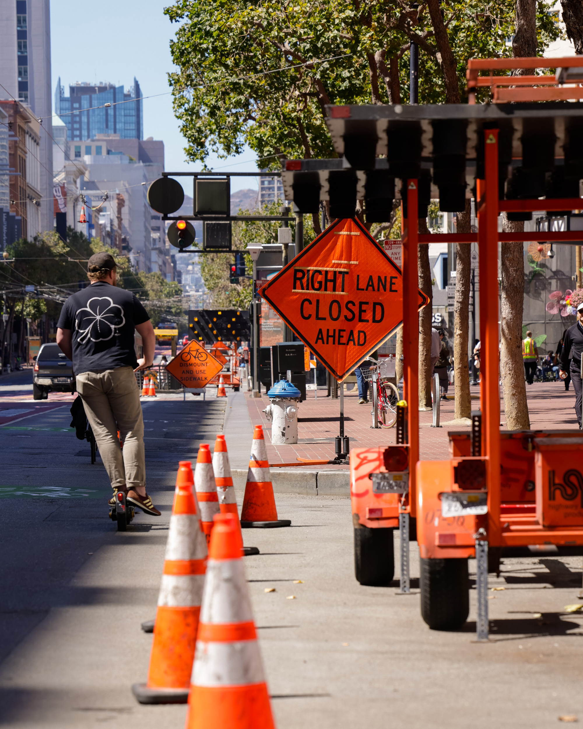 A person on a scooter travels along a city street lined with traffic cones and orange signs indicating a right lane closure ahead, under bright daylight.