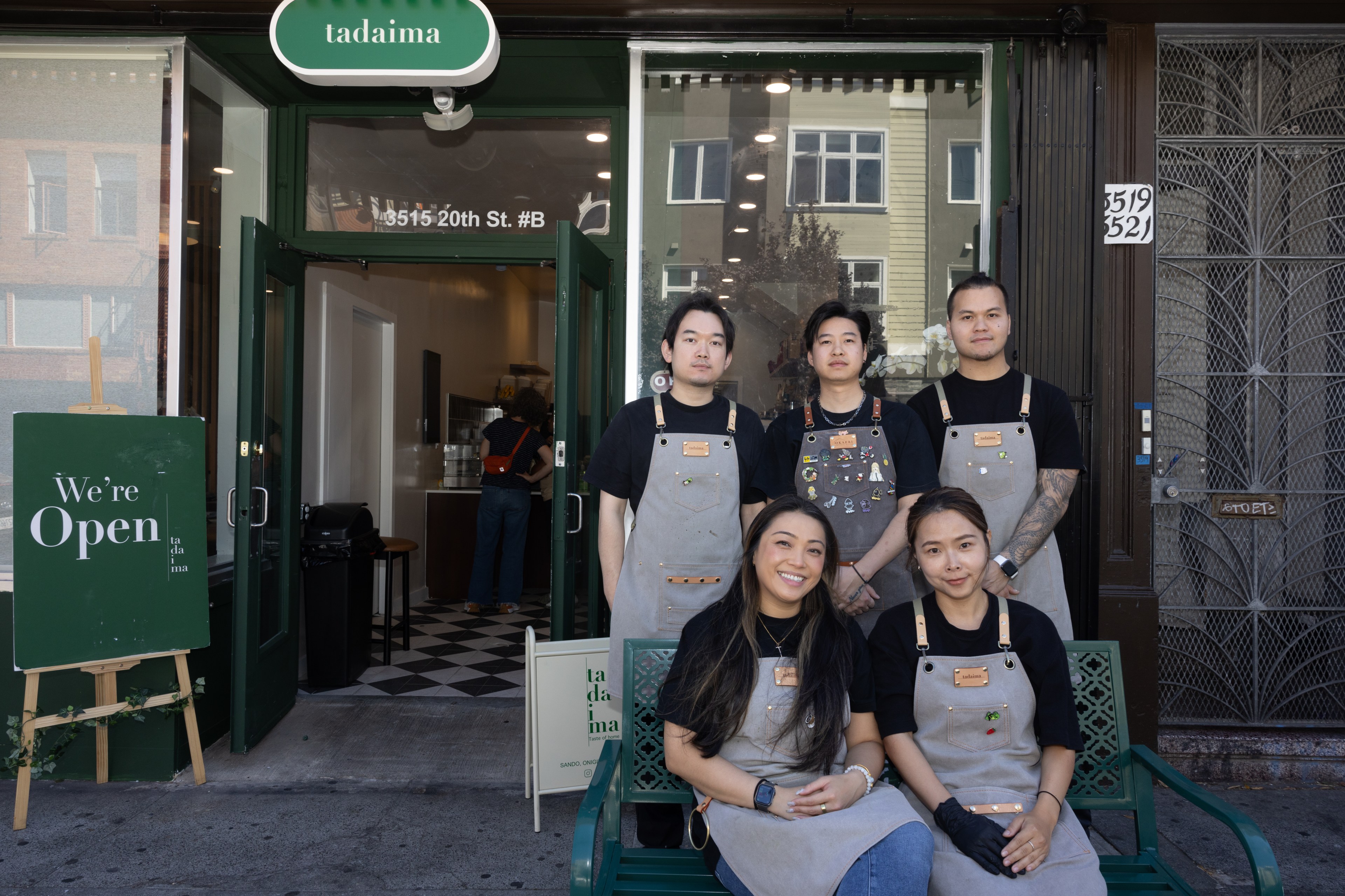 Five people in aprons stand and sit in front of a storefront named &quot;tadaima&quot; with a sign saying &quot;We're Open.&quot; The interior has checkered flooring.