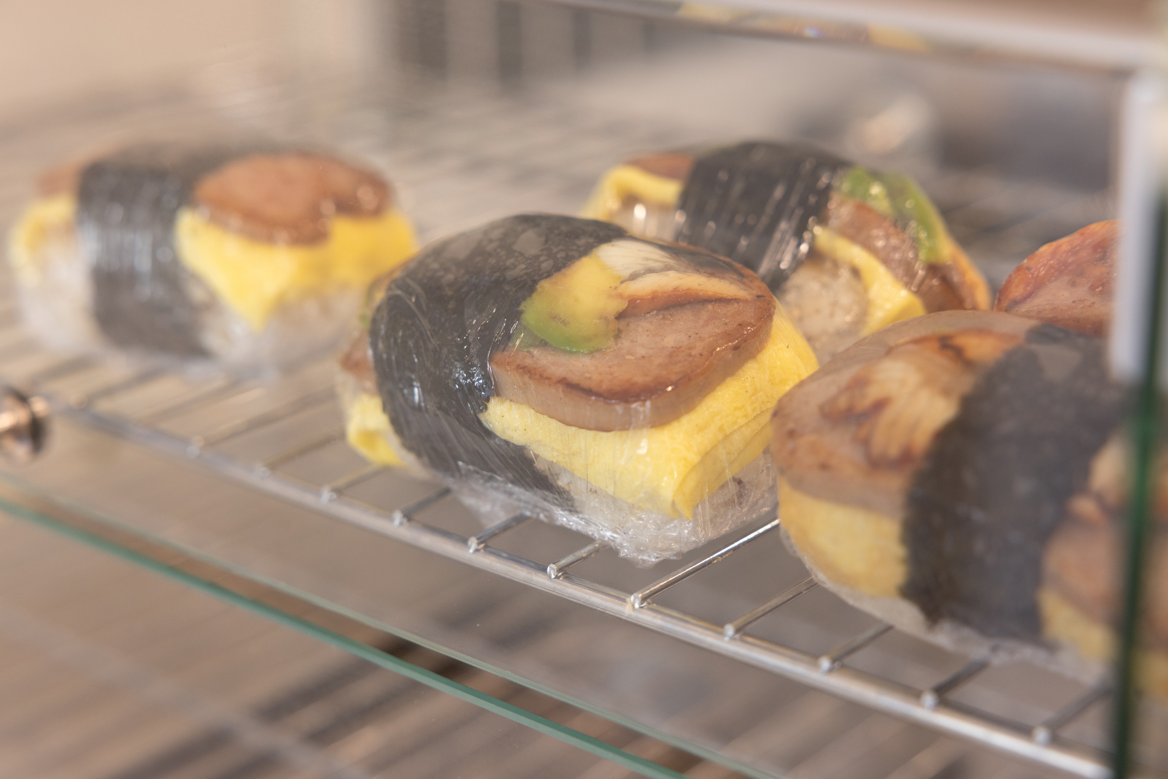 Four pieces of Spam musubi wrapped in plastic wrap sit on a metal rack in a display case. Each has rice, egg, and a slice of Spam, secured with seaweed.