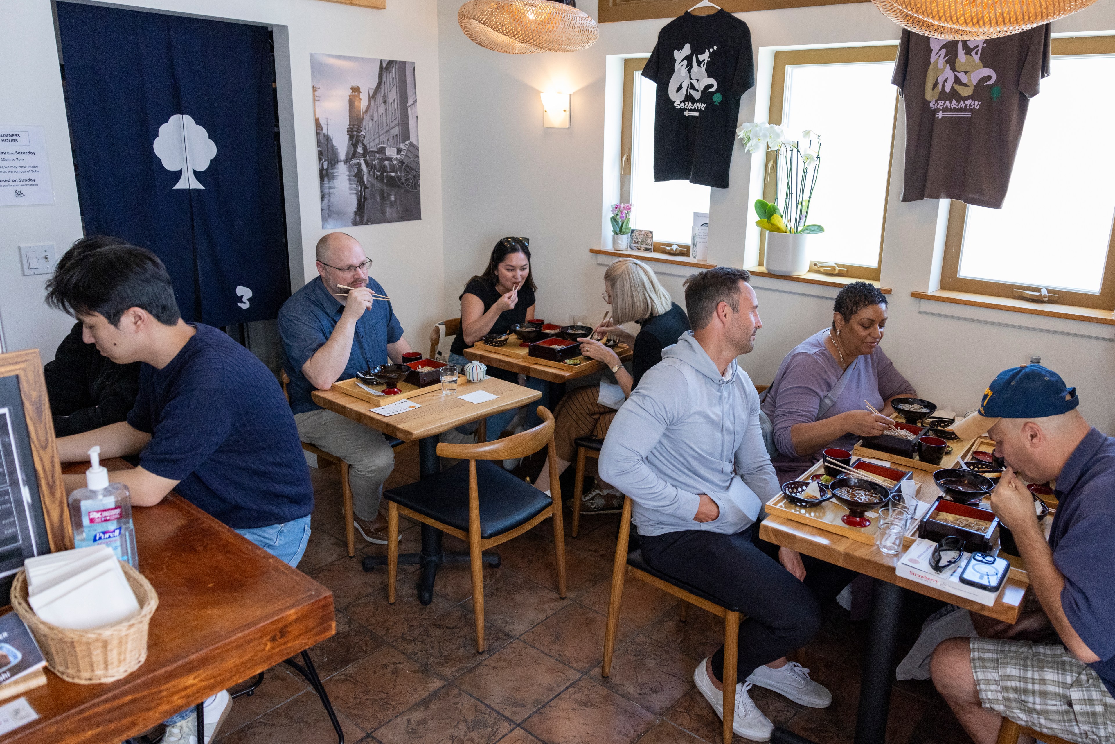 A cozy restaurant with several people seated at small wooden tables, enjoying meals. Two black shirts hang on the wall, and framed photos and decor adorn the space.