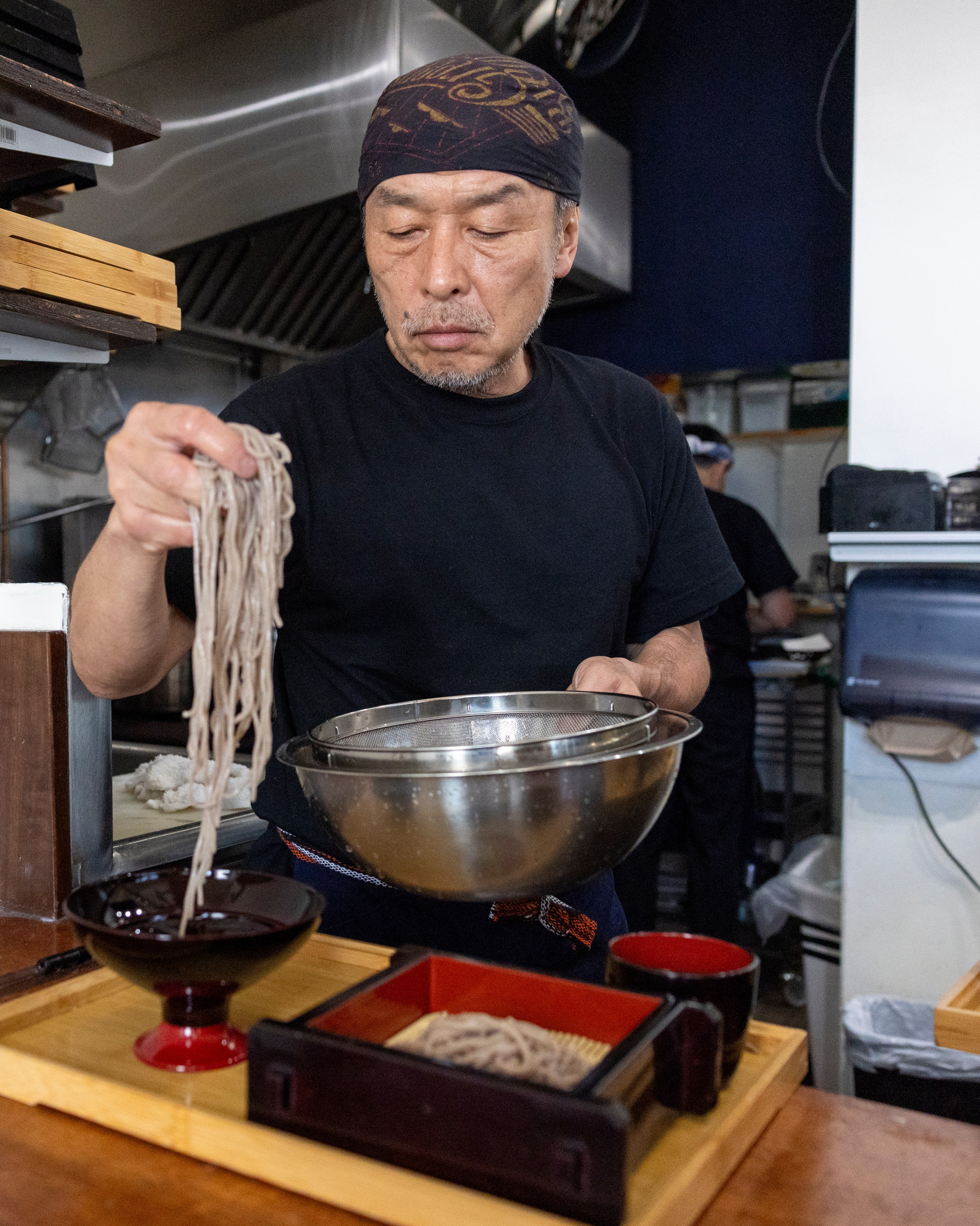 A man in a black shirt and bandana is preparing soba noodles in a kitchen, holding a metal strainer over a red bowl, with a wooden tray of noodles in front.