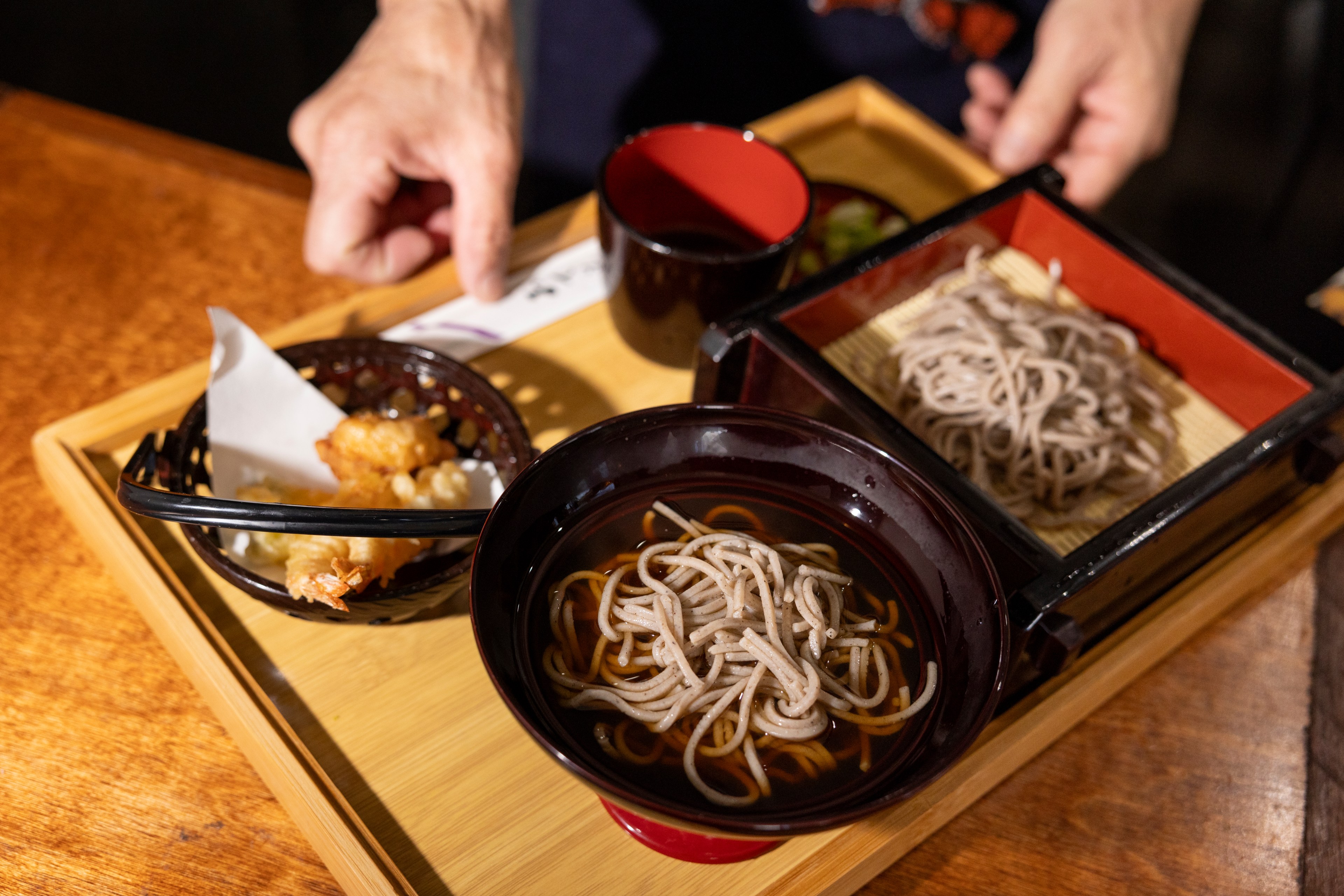 A tray holds a bowl of soba noodles in soup, a basket of tempura, and an additional serving of dry soba noodles. A hand is on the table, about to pick an item.