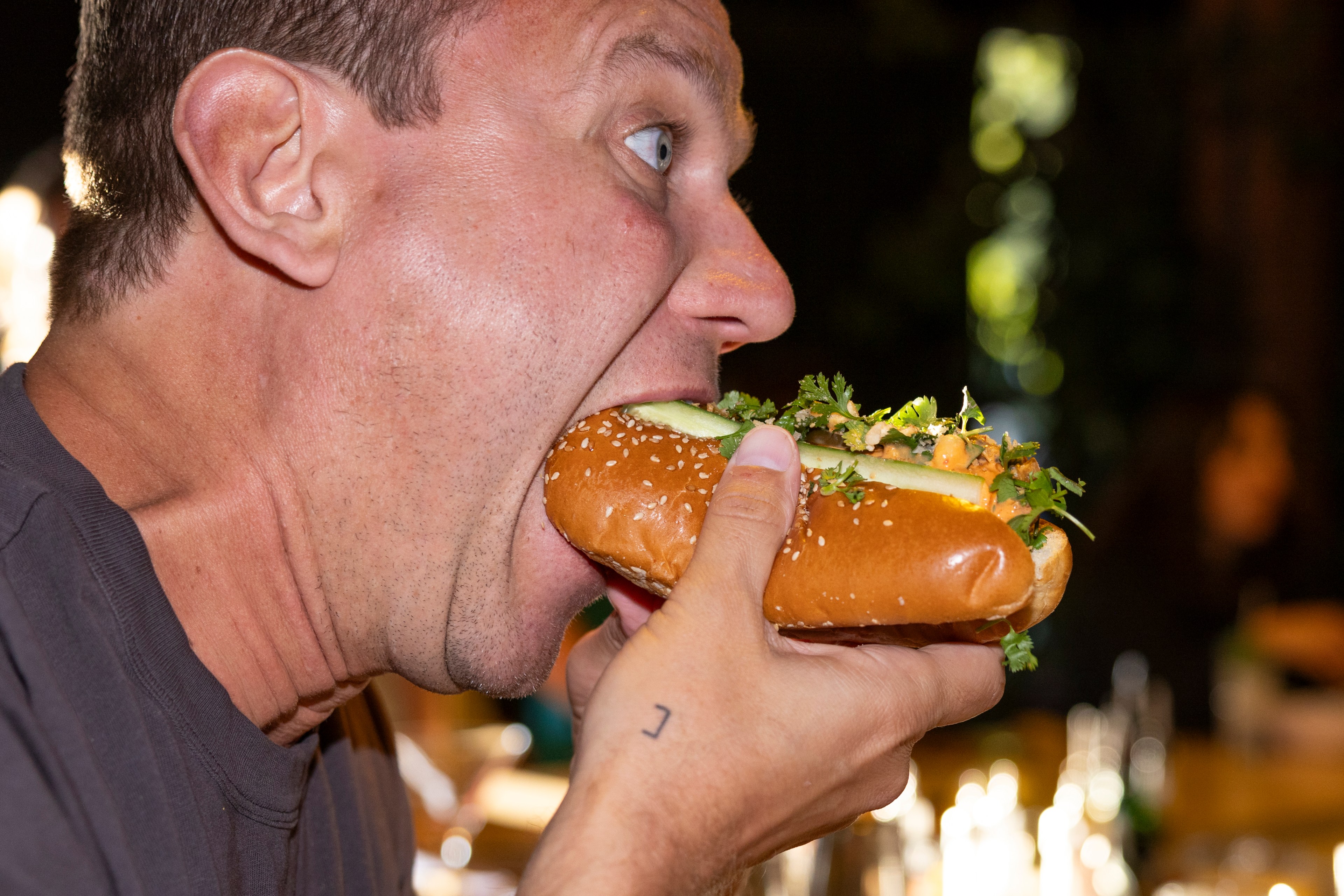 A man with a shocked expression is taking a large bite of a loaded sandwich on a sesame seed bun.