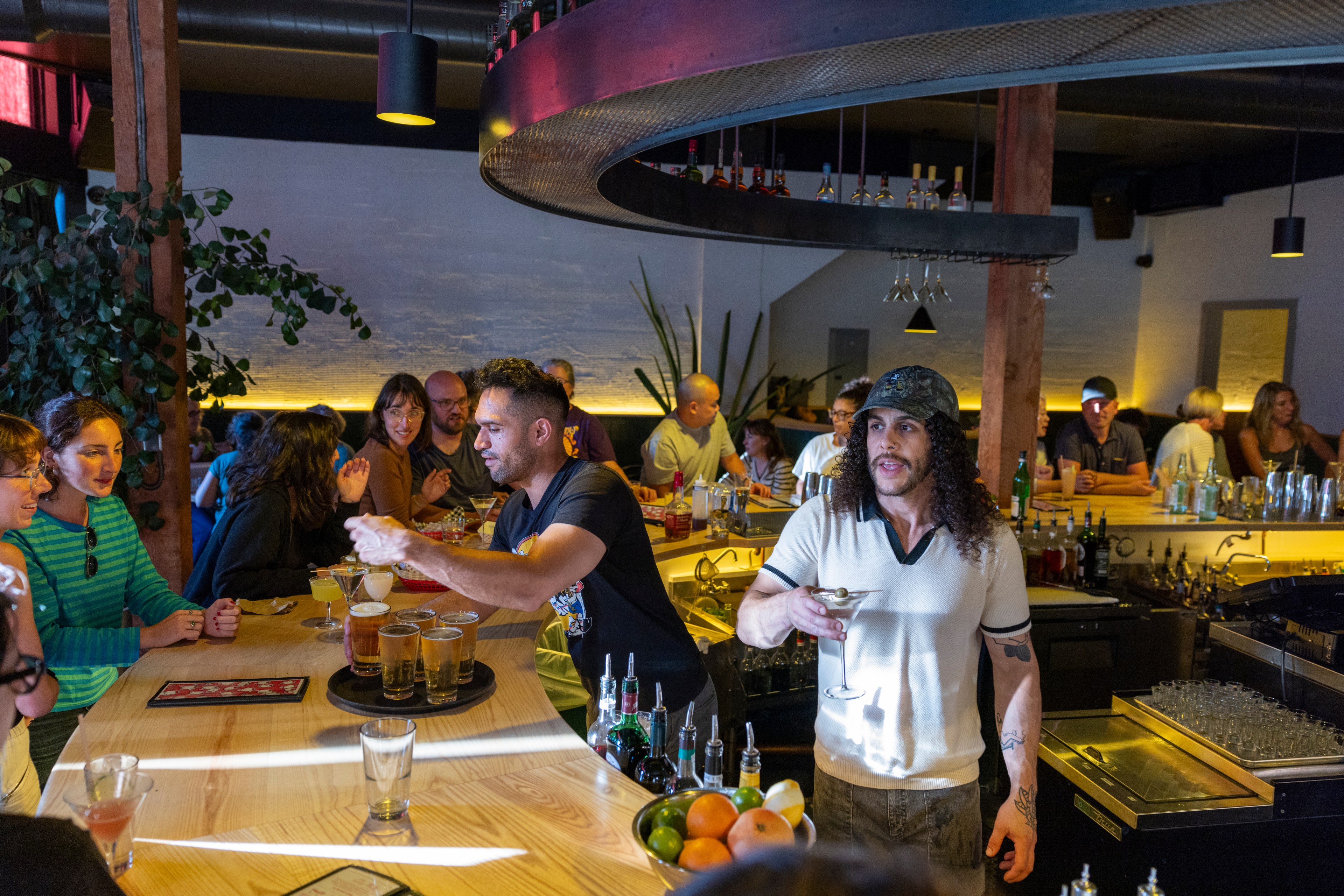 A lively bar scene features patrons conversing and enjoying drinks, while bartenders serve various beverages. The atmosphere is warm with modern industrial décor.