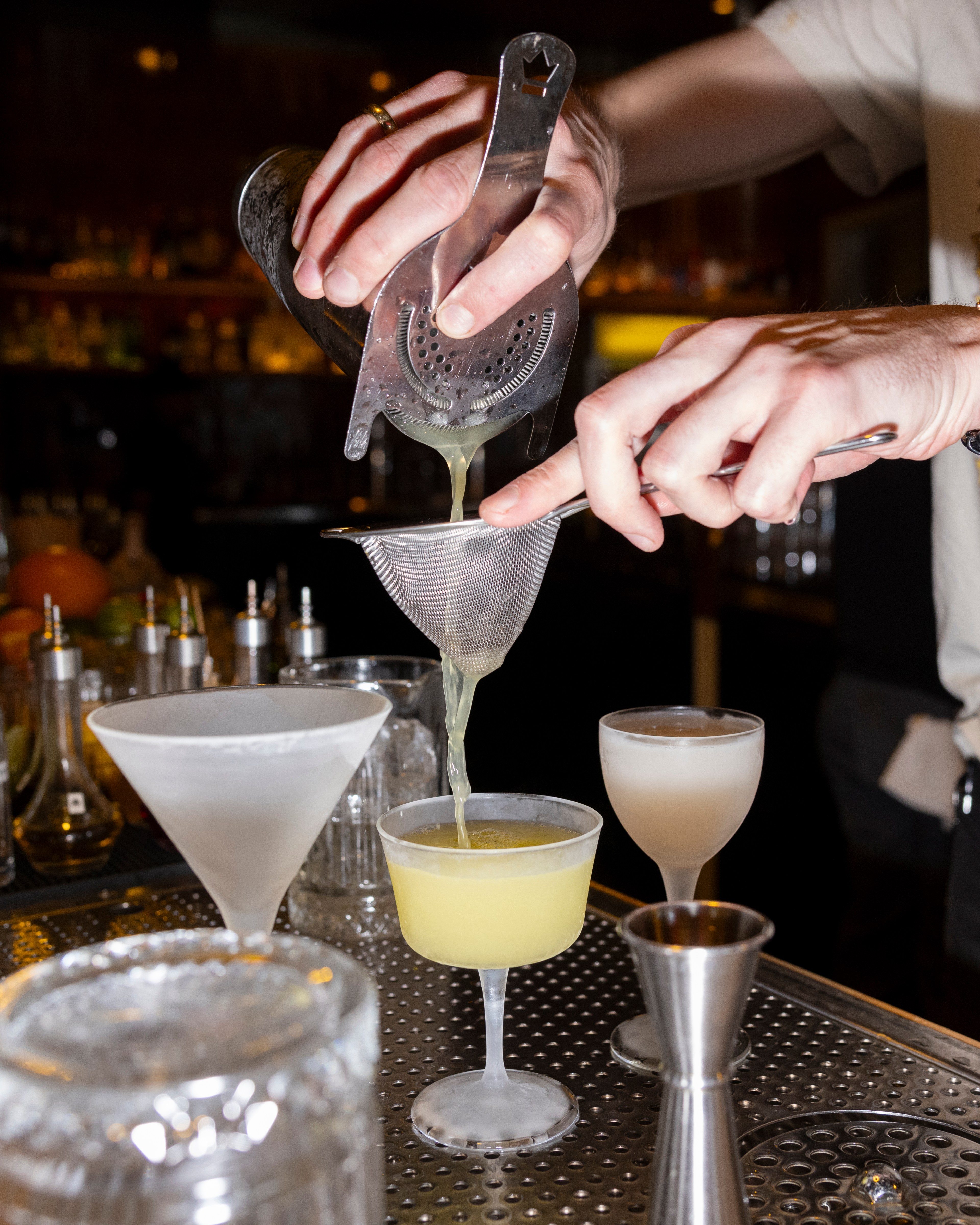 A pair of hands pours a cocktail mixture through a strainer into a frosted glass. Two other frosted glasses, one white and one yellow, are on the bar counter.