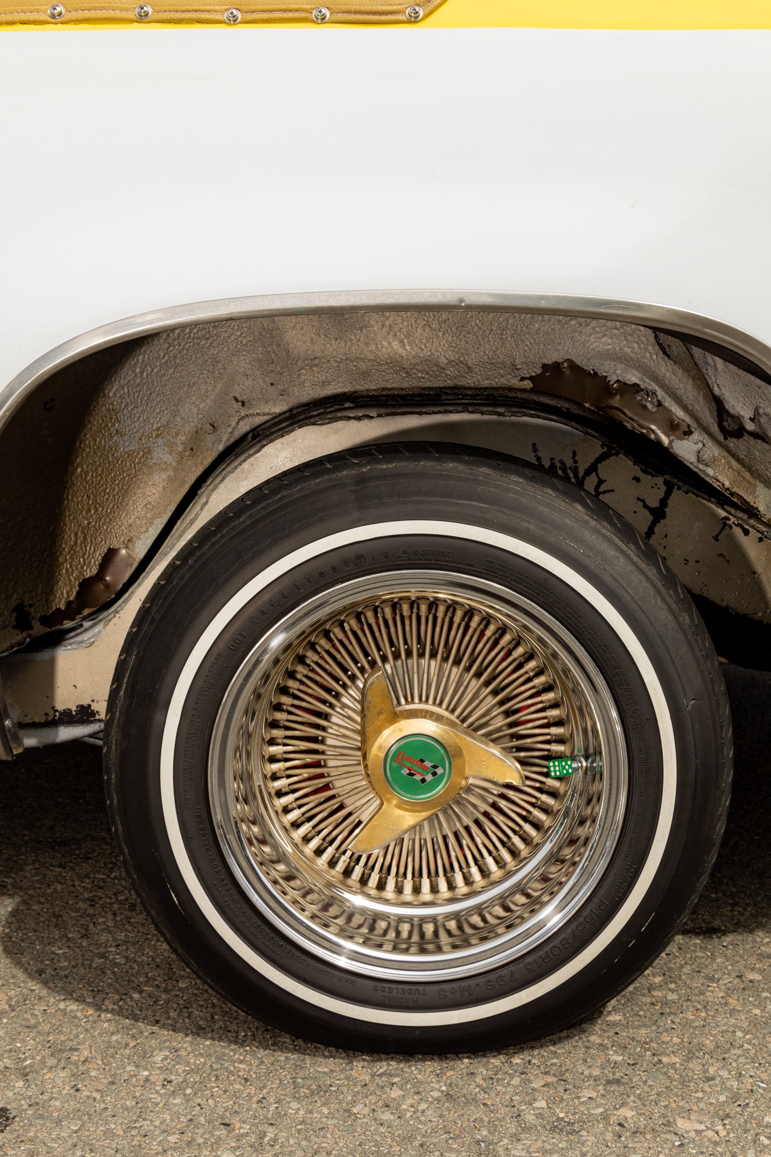 The image shows a close-up of a vintage car's rear wheel with a gold wire-spoke rim, whitewall tire, and a white and yellow paint job. The car's wheel well appears slightly rusted.