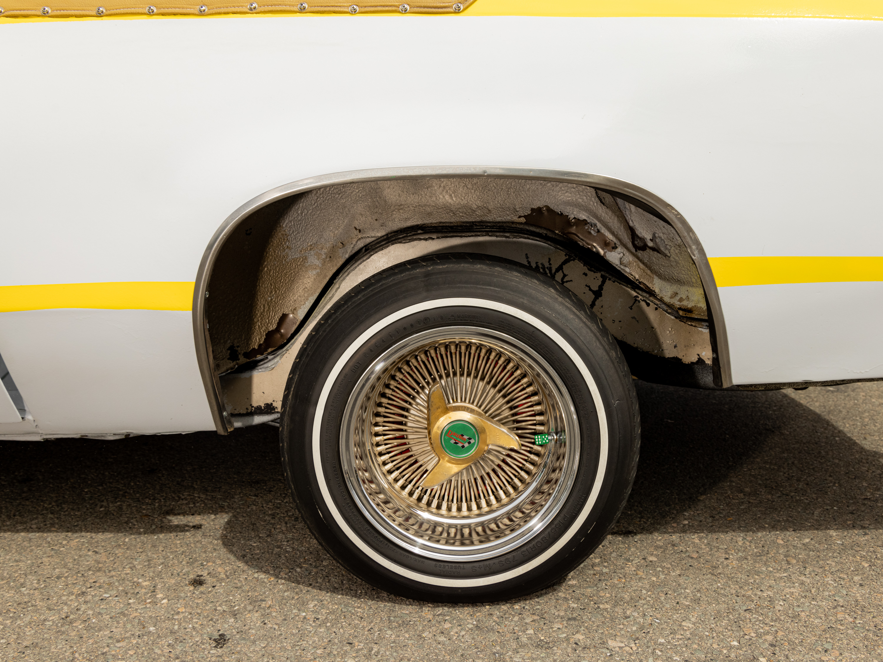 The image shows a close-up of a vintage car's rear wheel with a gold wire-spoke rim, whitewall tire, and a white and yellow paint job. The car's wheel well appears slightly rusted.