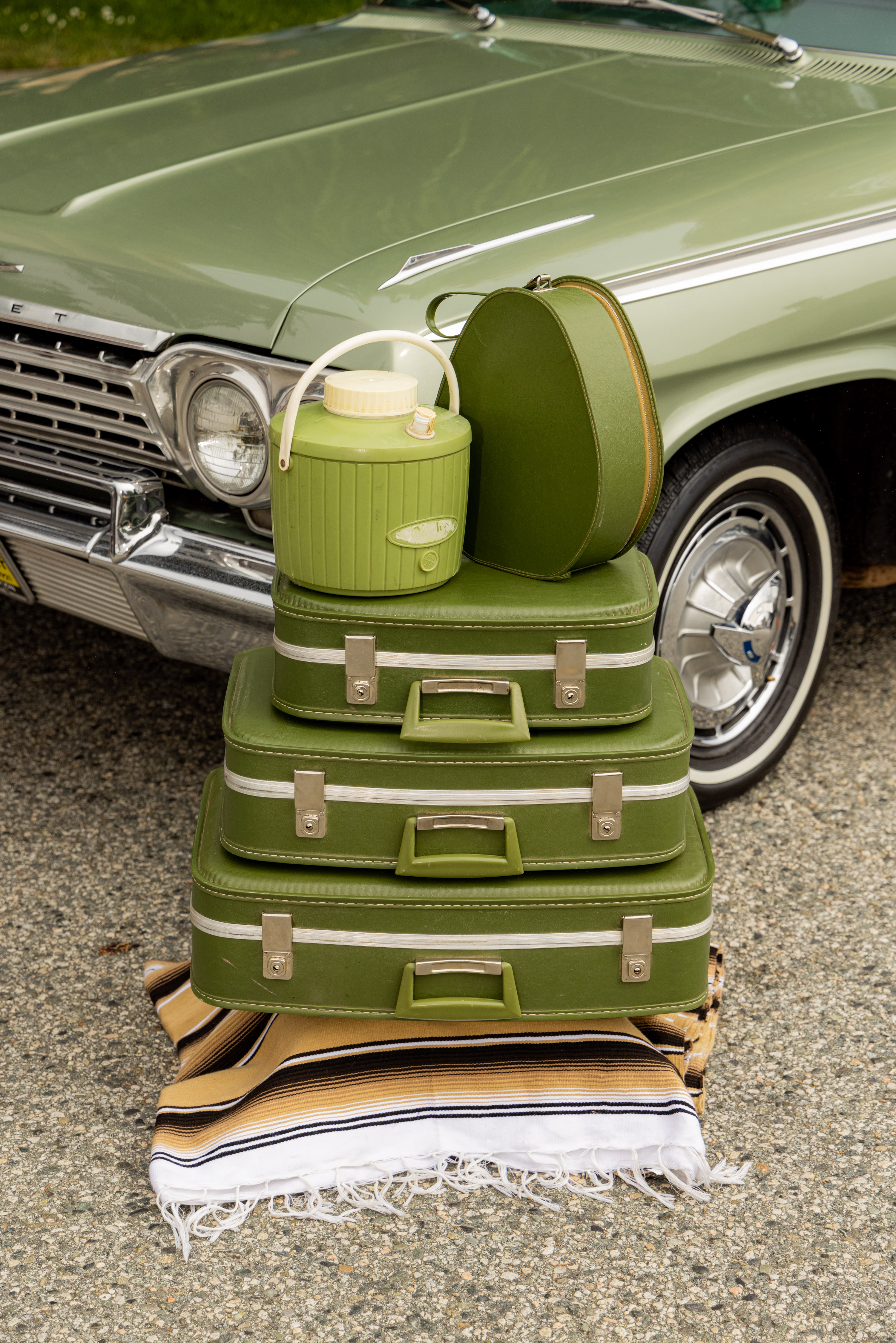 A stack of green vintage suitcases, a round hatbox, and a green water jug sit on a striped blanket in front of a classic green car.