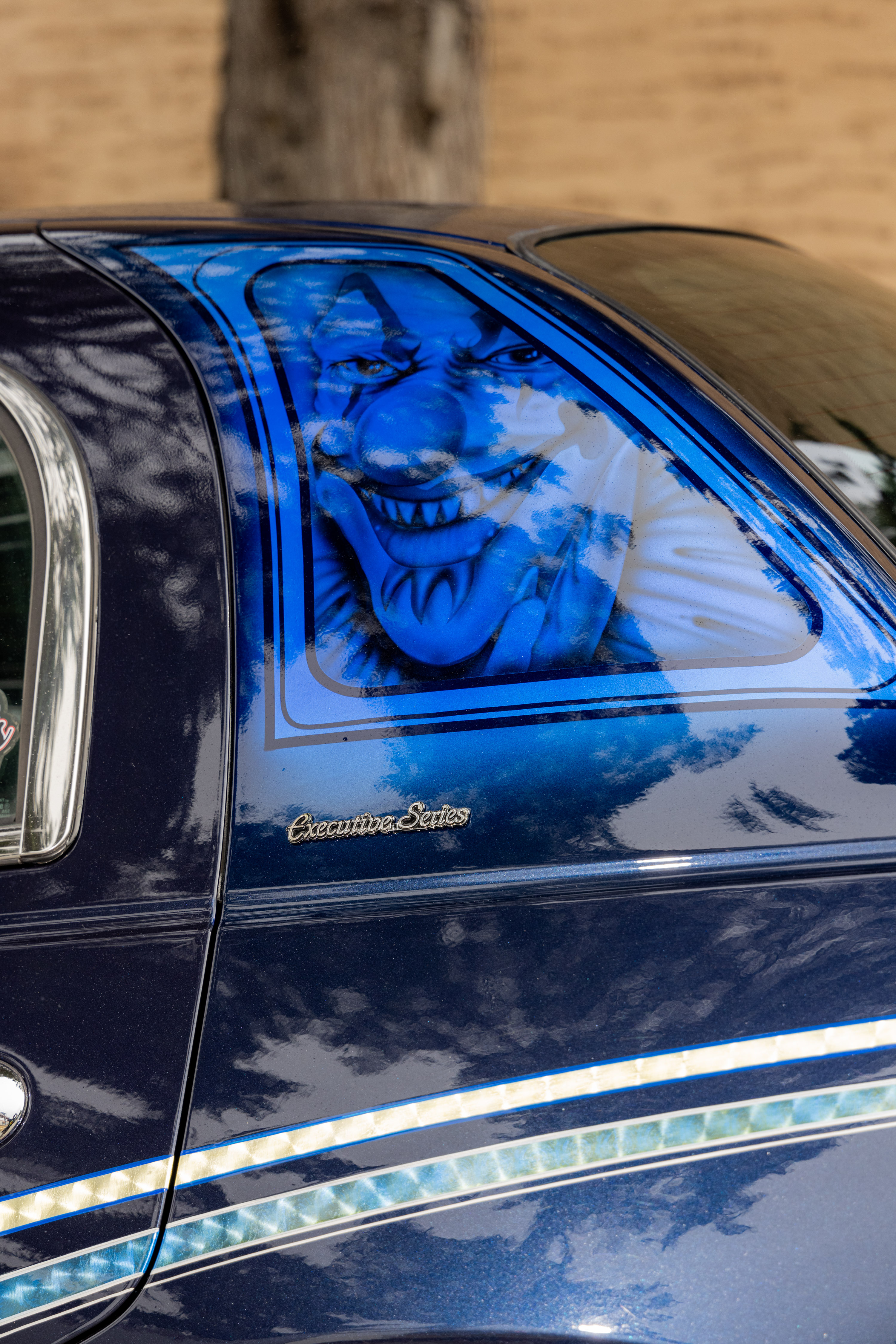 The image shows a dark blue car with a menacing blue clown face painted on the rear side panel. It has &quot;Executive Series&quot; written below the painting.