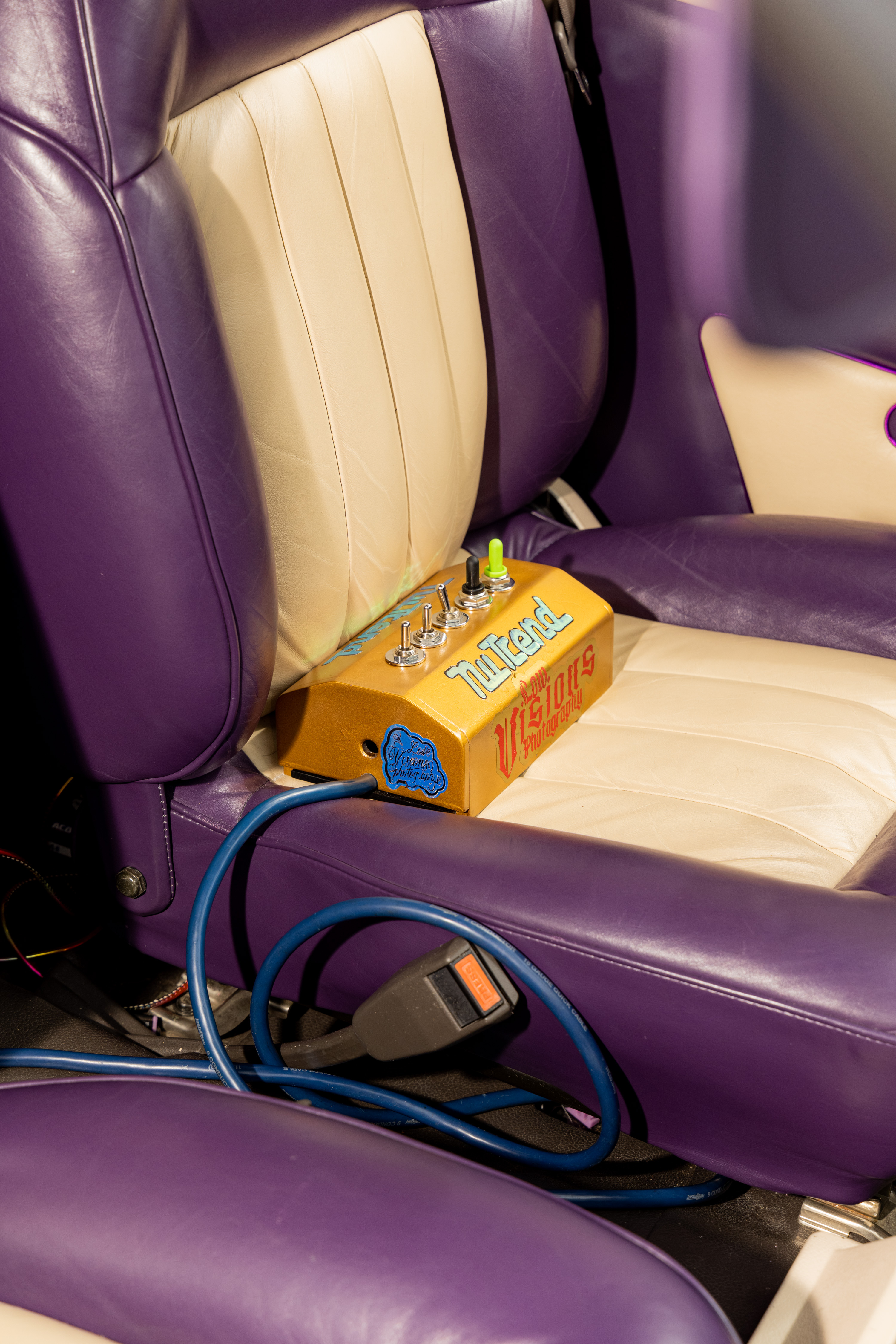 A gold metal box with switches and colorful labels sits on a cream and purple leather car seat, with a blue cable connected to it and a visible seatbelt buckle nearby.