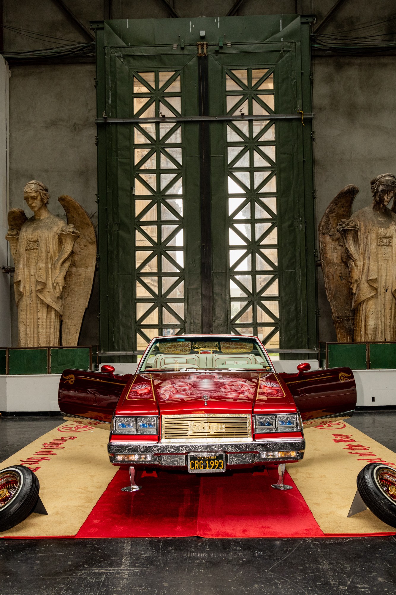 The image shows a vintage car exhibition with a red convertible in the center on a carpet, flanked by white and mint-green cars. Two large angel statues are in the background.