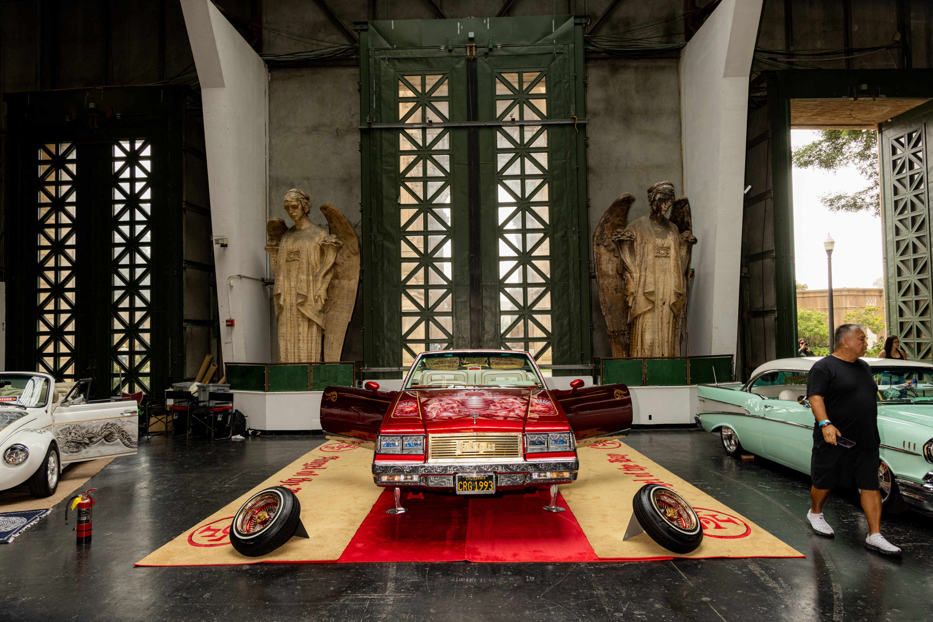 The image shows a vintage car exhibition with a red convertible in the center on a carpet, flanked by white and mint-green cars. Two large angel statues are in the background.