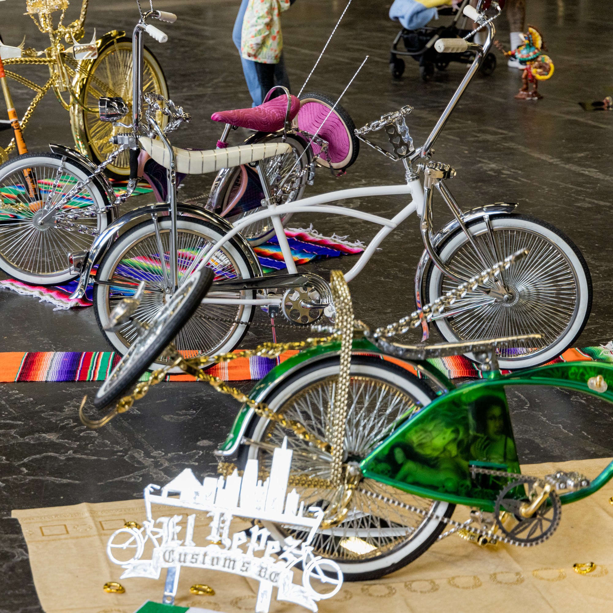 The image shows a display of customized lowrider bicycles with elaborate designs and colorful accessories, set on vibrant blankets in an indoor exhibition space.