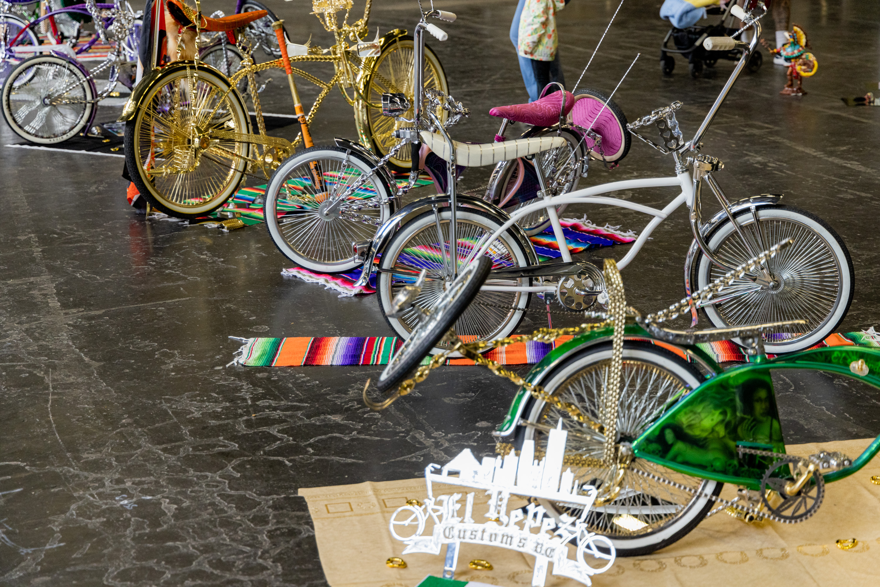 The image shows a display of customized lowrider bicycles with elaborate designs and colorful accessories, set on vibrant blankets in an indoor exhibition space.