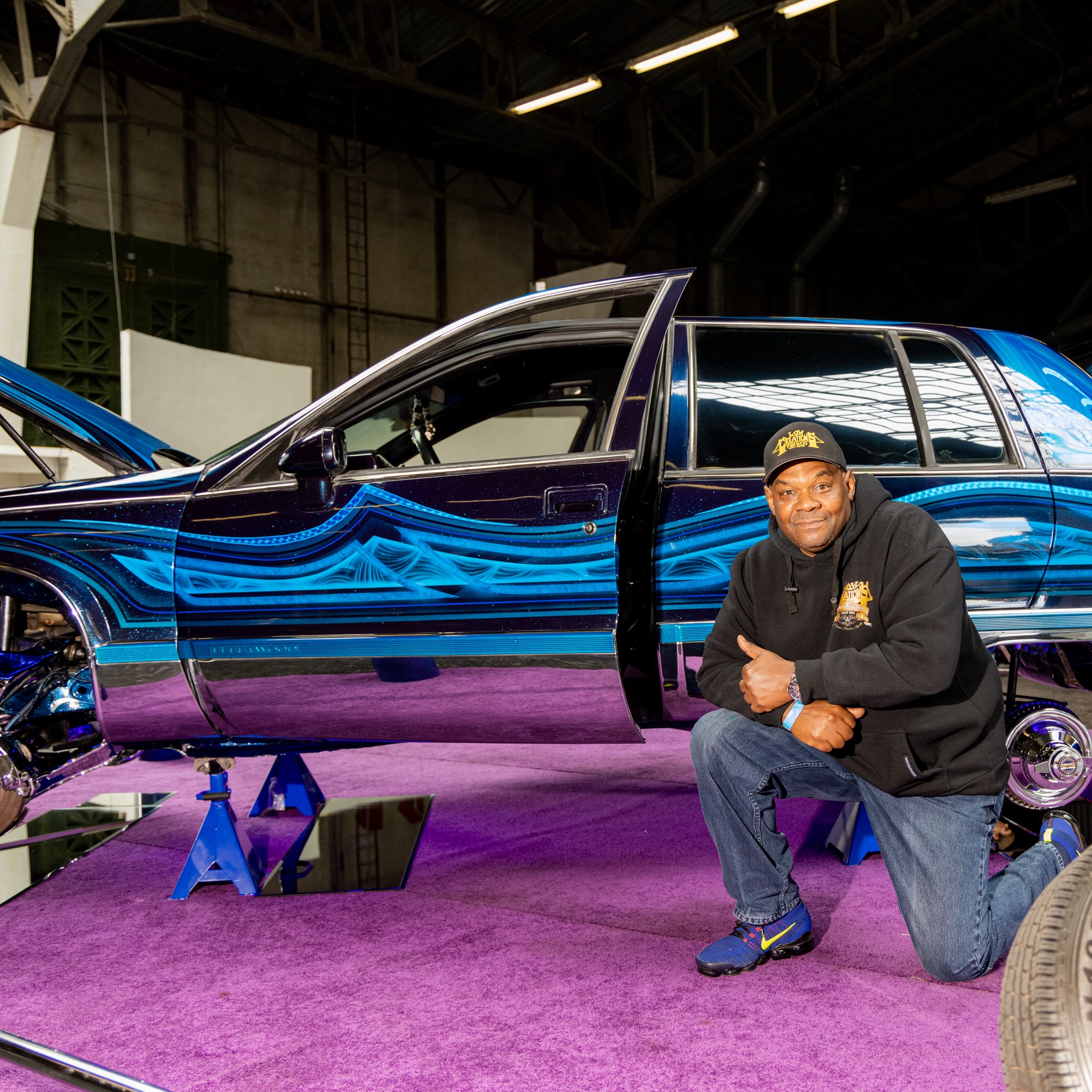 A man kneels on a purple carpet beside a custom car with intricate blue patterns and opened hood. The car is lifted on stands, showcasing chrome details.