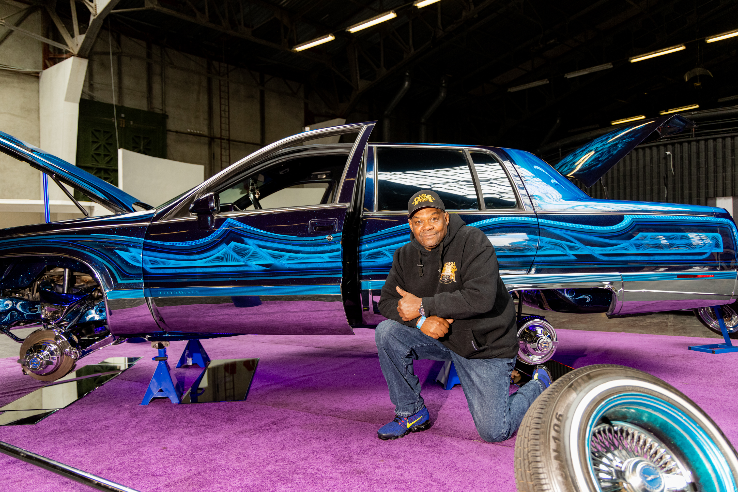 A man kneels on a purple carpet beside a custom car with intricate blue patterns and opened hood. The car is lifted on stands, showcasing chrome details.