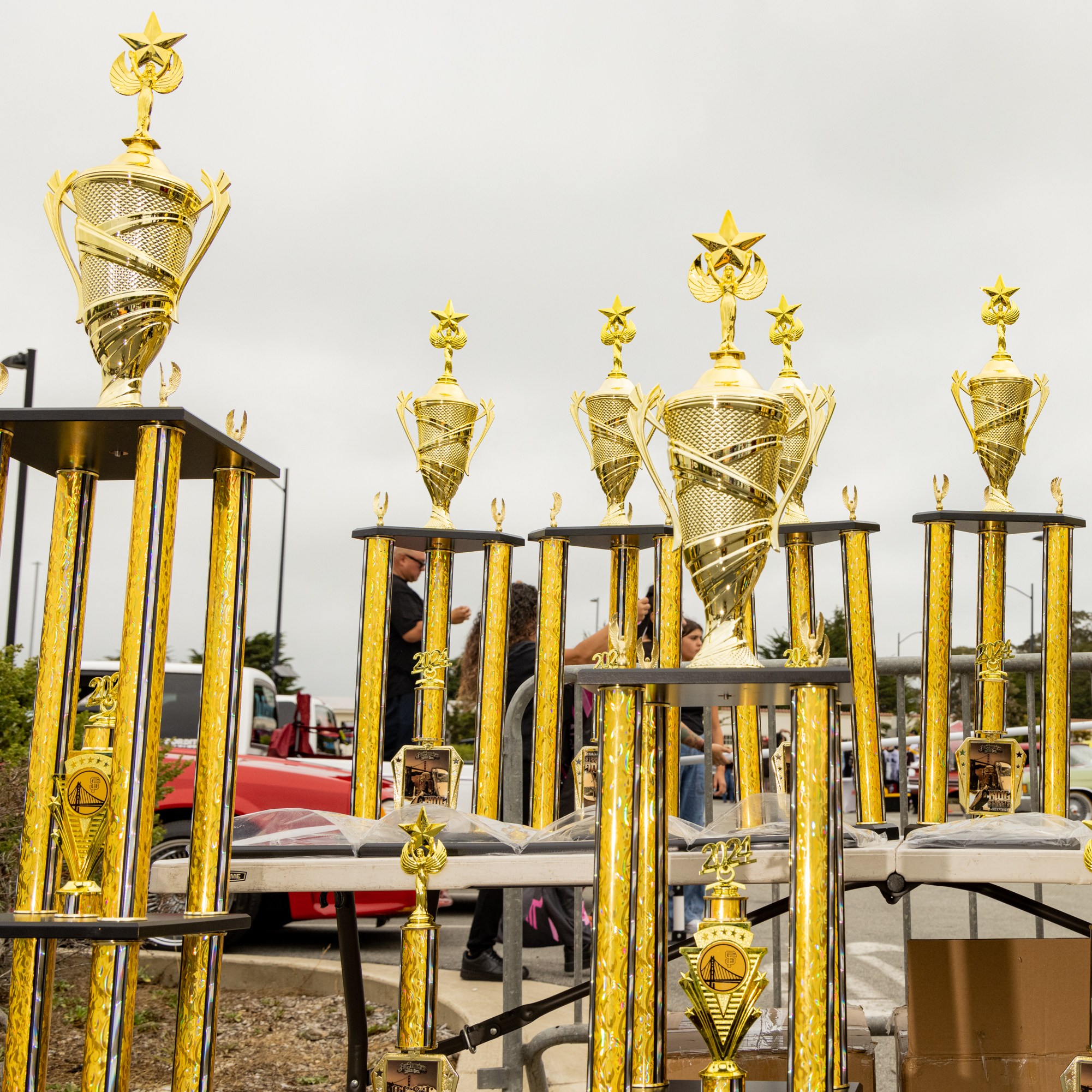 The image shows a variety of tall, golden trophies with intricate designs and star tops, displayed on tables outdoors with a cloudy sky in the background.